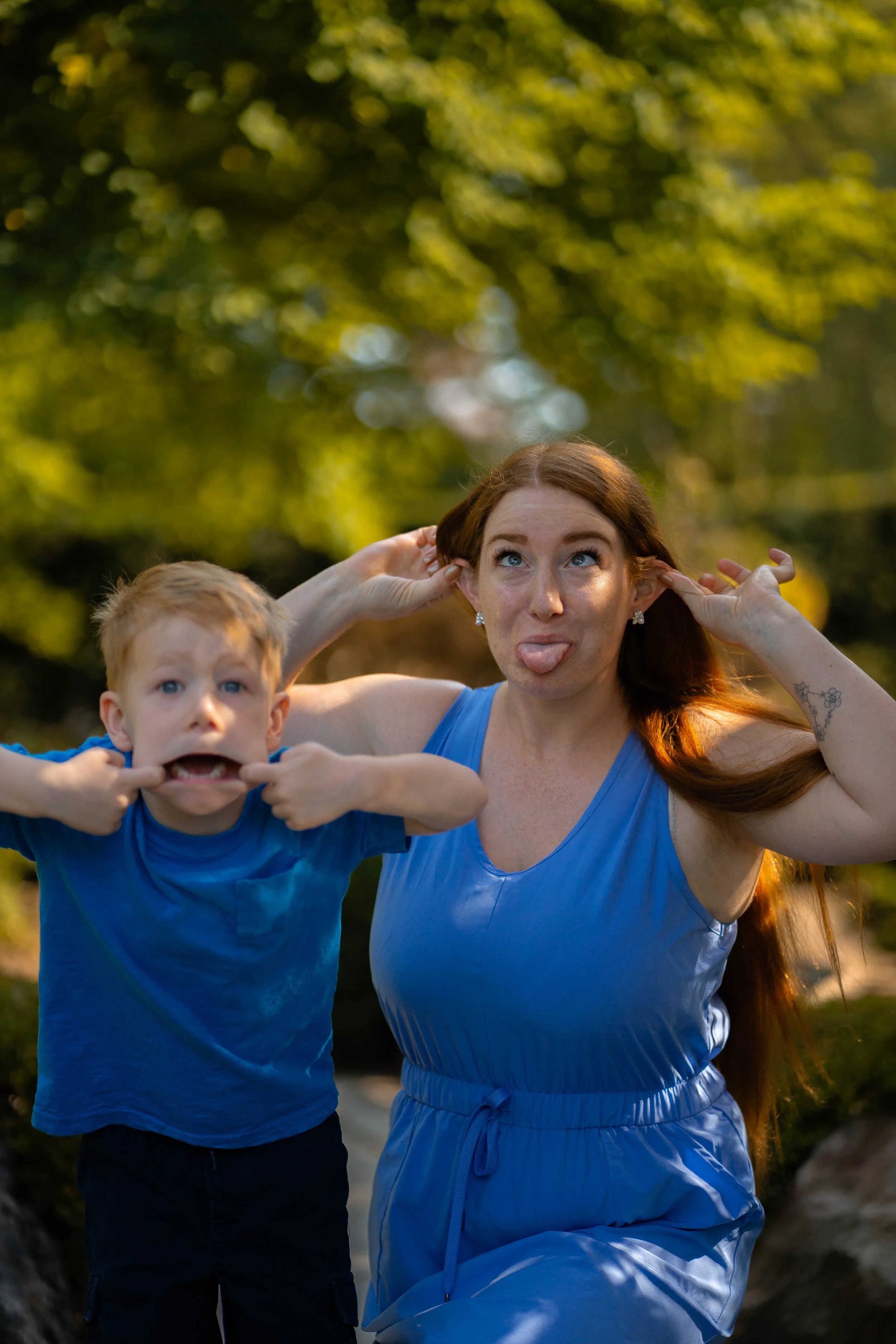 A woman and a boy making funny faces outdoors with green trees in the background.