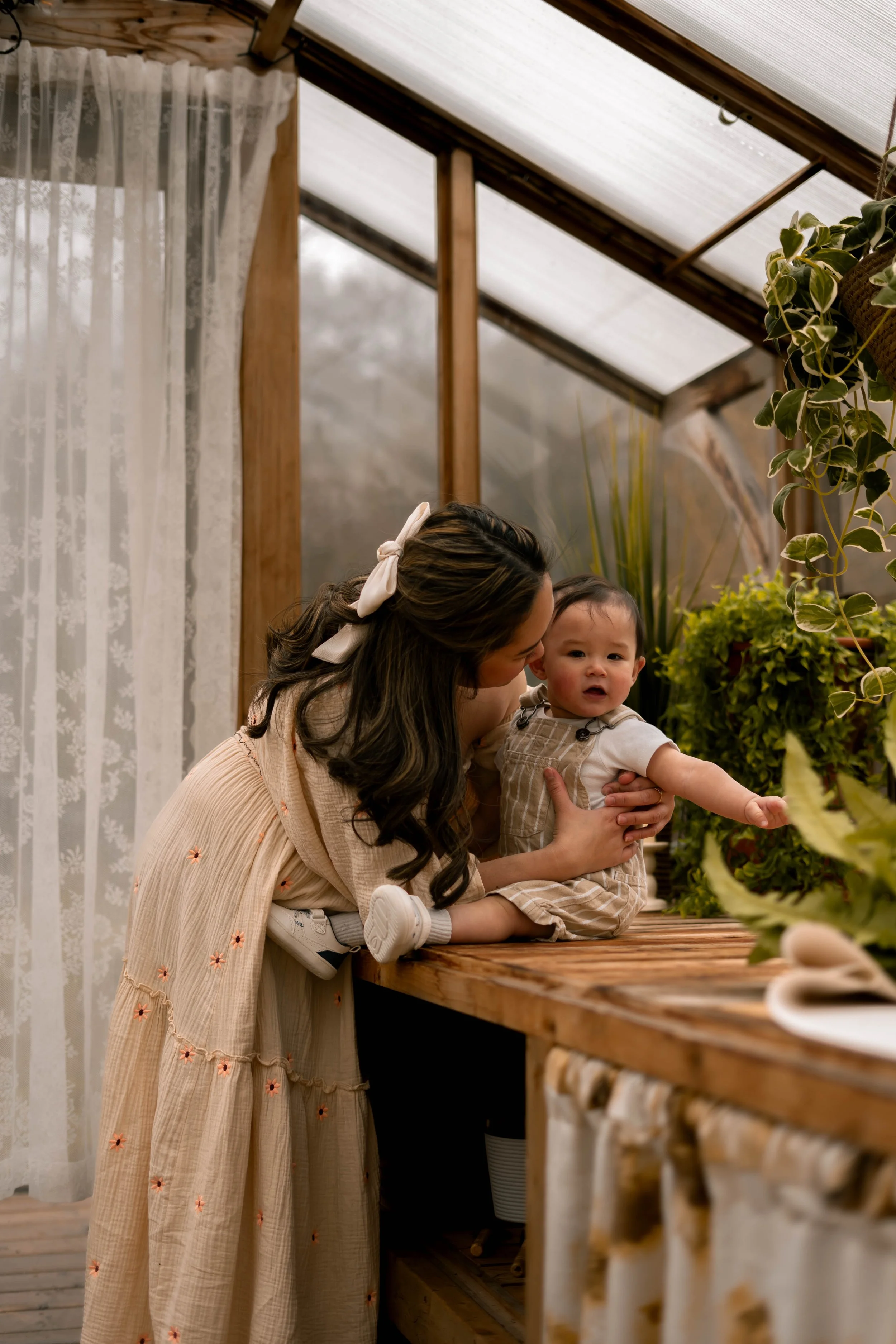A woman with dark wavy hair and a cream-colored dress with orange embroidery is kneeling on a wooden surface, holding a young child with dark hair, wearing a plaid shirt and beige overalls. They are inside a warmly lit room with wooden beams, potted 