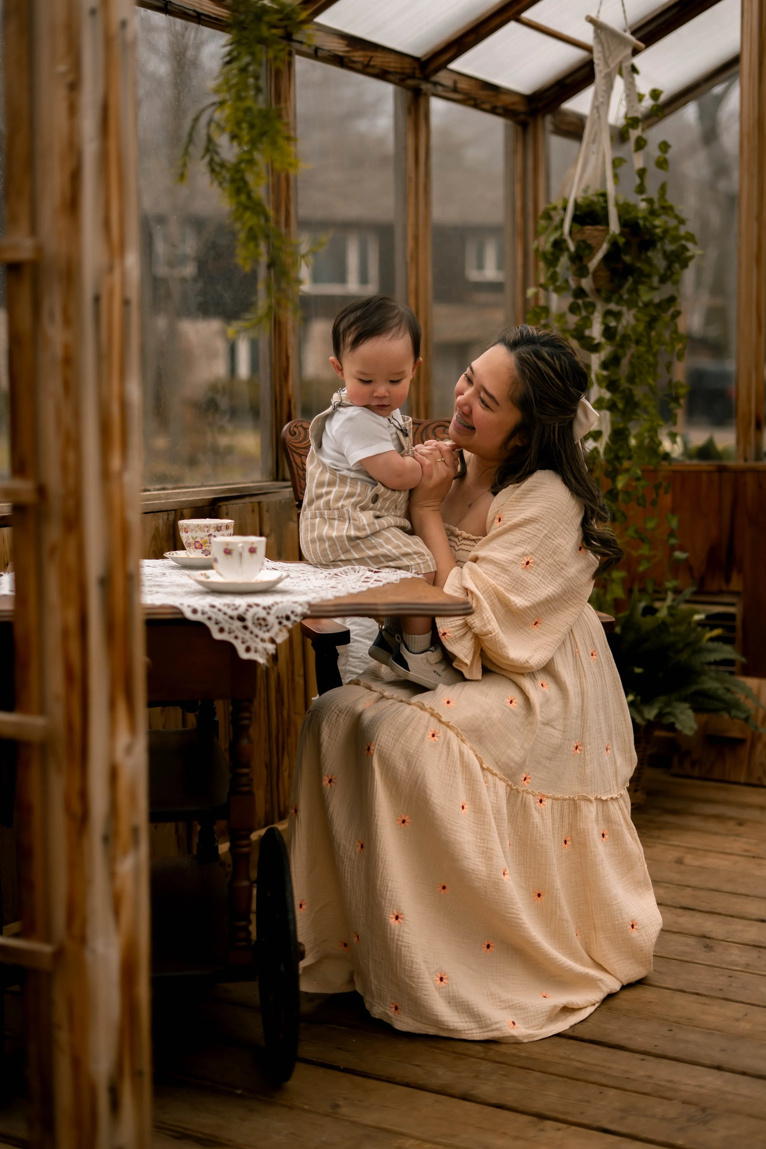 A woman with dark hair in a light beige dress with embroidered flowers is sitting on the floor, holding a young child on her lap. The child is wearing a striped outfit. They are inside a wooden and glass greenhouse with hanging plants and a wooden ta