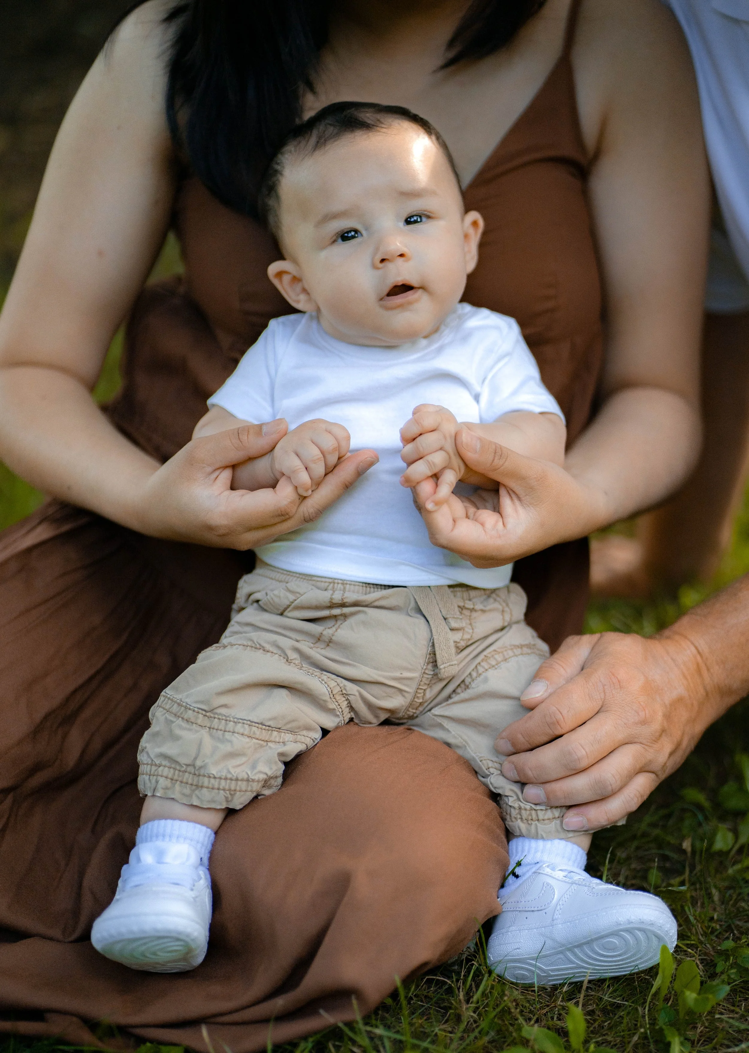 A baby sitting on an adult's lap outdoors, holding hands with a person behind them. The baby is wearing a white t-shirt, beige pants, and white sneakers, and has short dark hair. The adult, partially visible, is wearing a brown dress. The setting is
