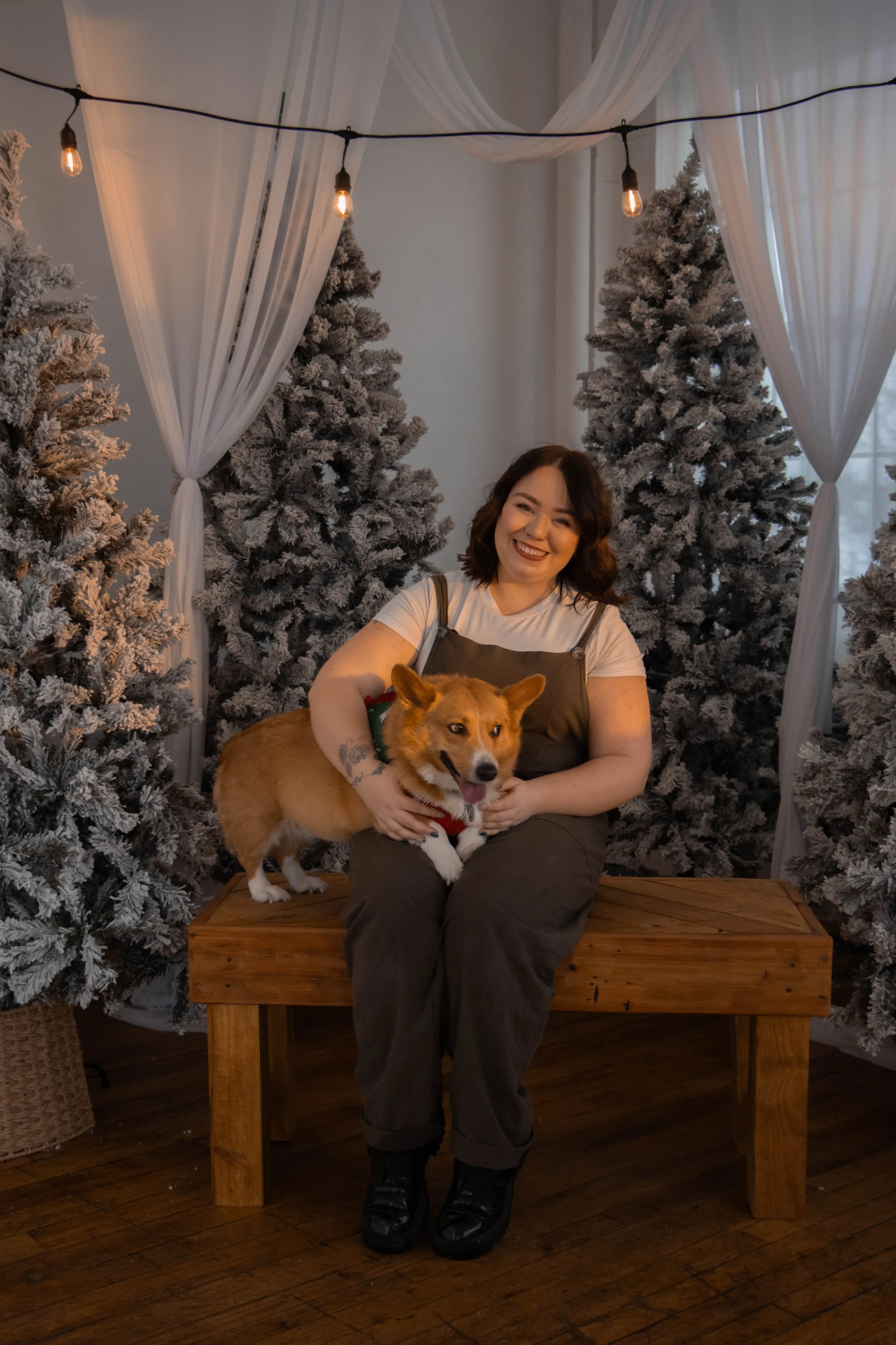 A woman with dark hair sitting on a wooden bench, smiling, holding a dog with a red collar. The background features several snow-flocked Christmas trees, wrapped in white curtains and hanging string lights.