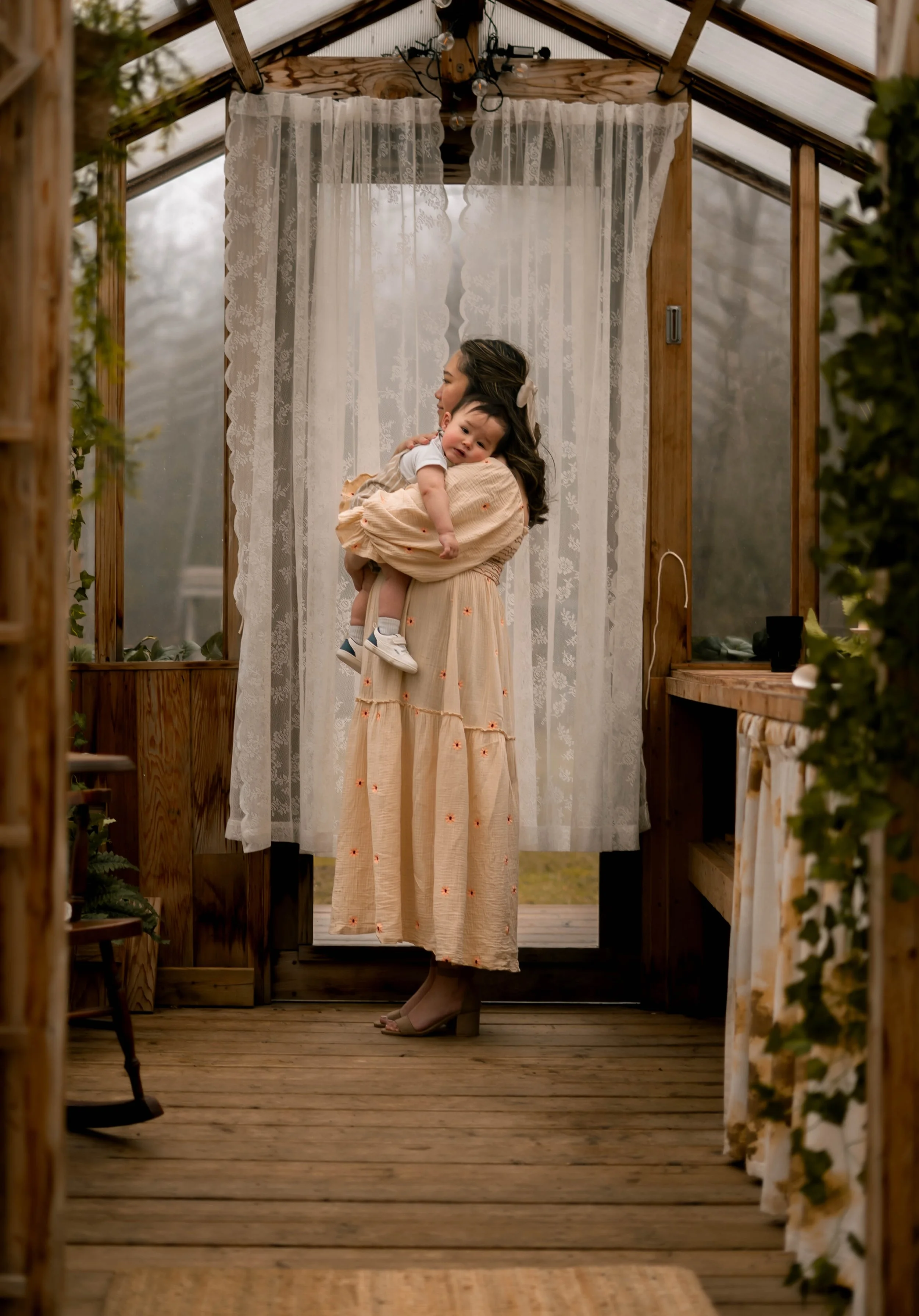 A woman holding a young child in a wooden greenhouse or conservatory with sheer lace curtains, wooden frames, and string lights.