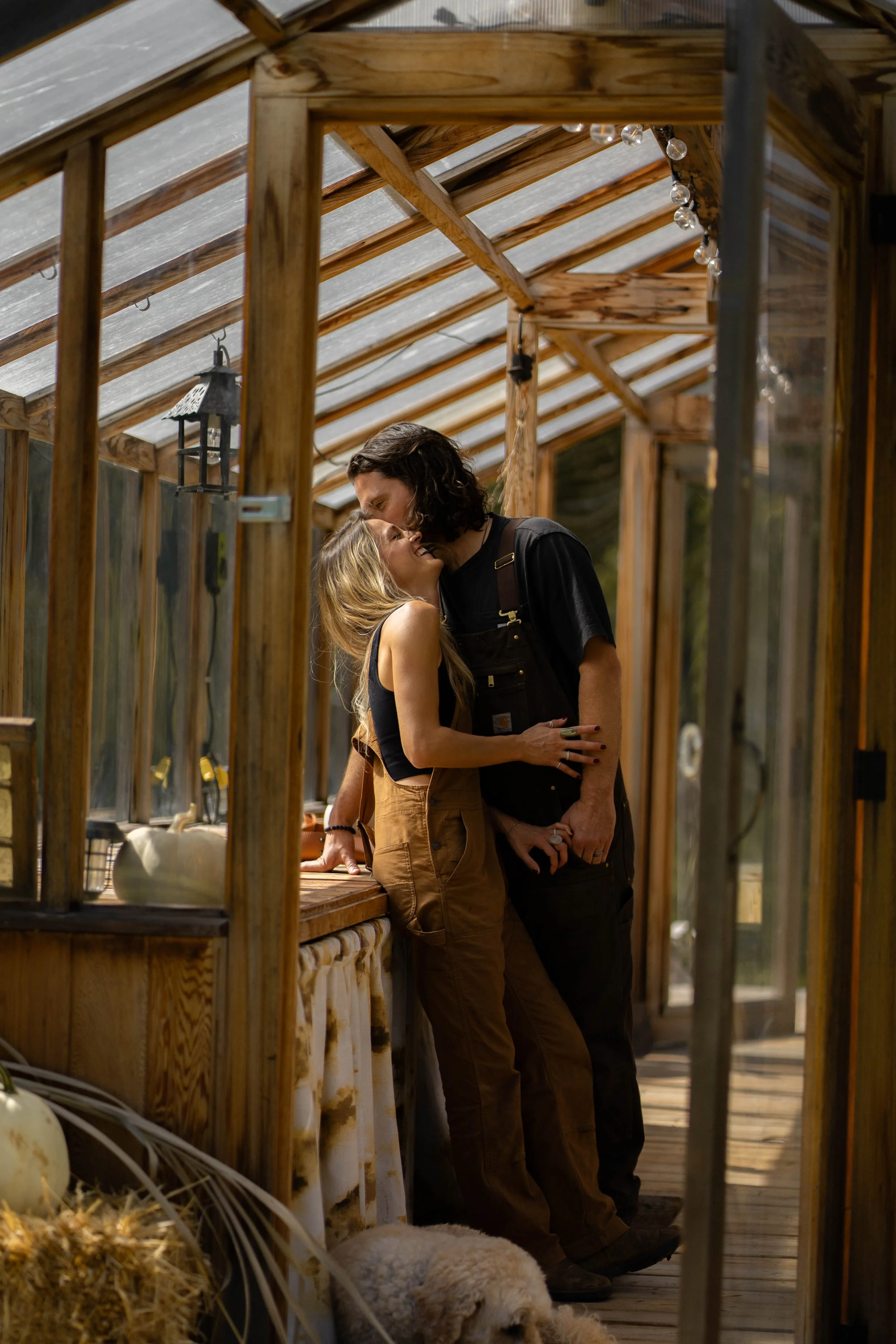 A family of four standing in front of a fountain in a park on a sunny day. The father and mother are smiling at each other, with the mother holding a young girl and the father holding a young boy. The girl has red hair and wears a light-colored dress, while the boy has red hair and wears a blue shirt and shorts.