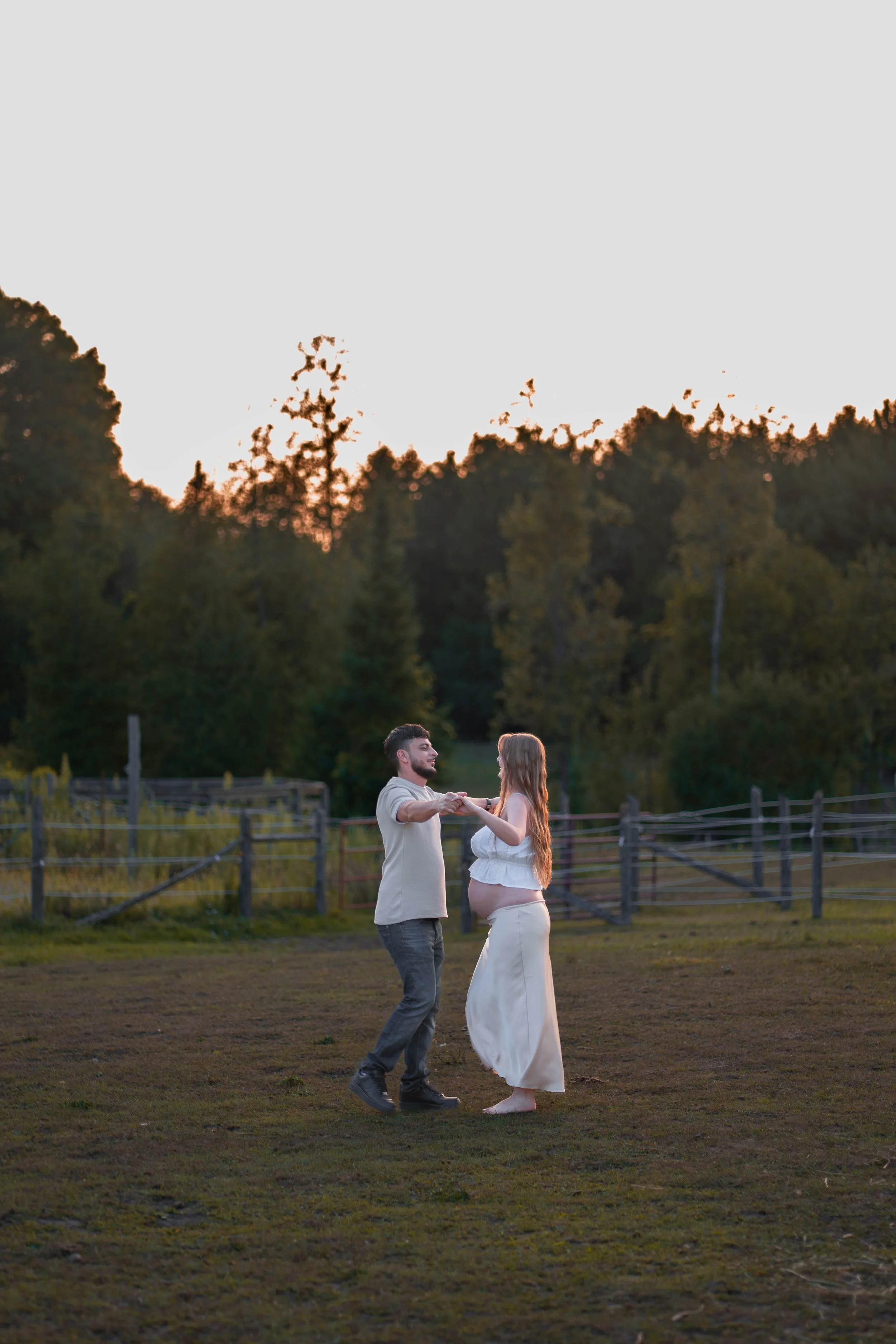 A couple dancing outdoors on a grassy field at sunset, with a wooden fence and trees in the background.
