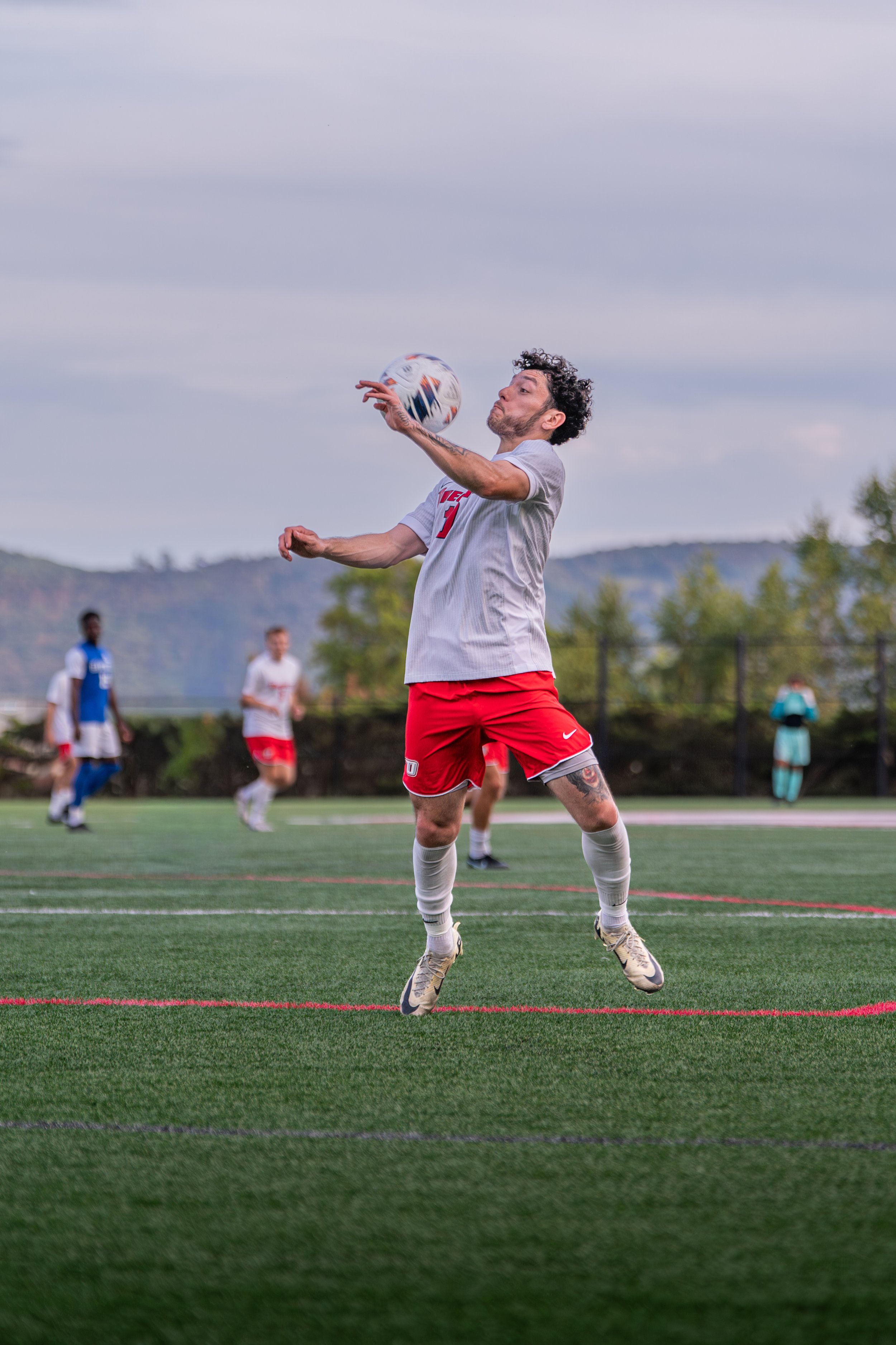 A male soccer player in a gray shirt and red shorts is jumping in the air, reaching to catch a soccer ball during a game on a green field. Other players and a person in a teal outfit are visible in the background.
