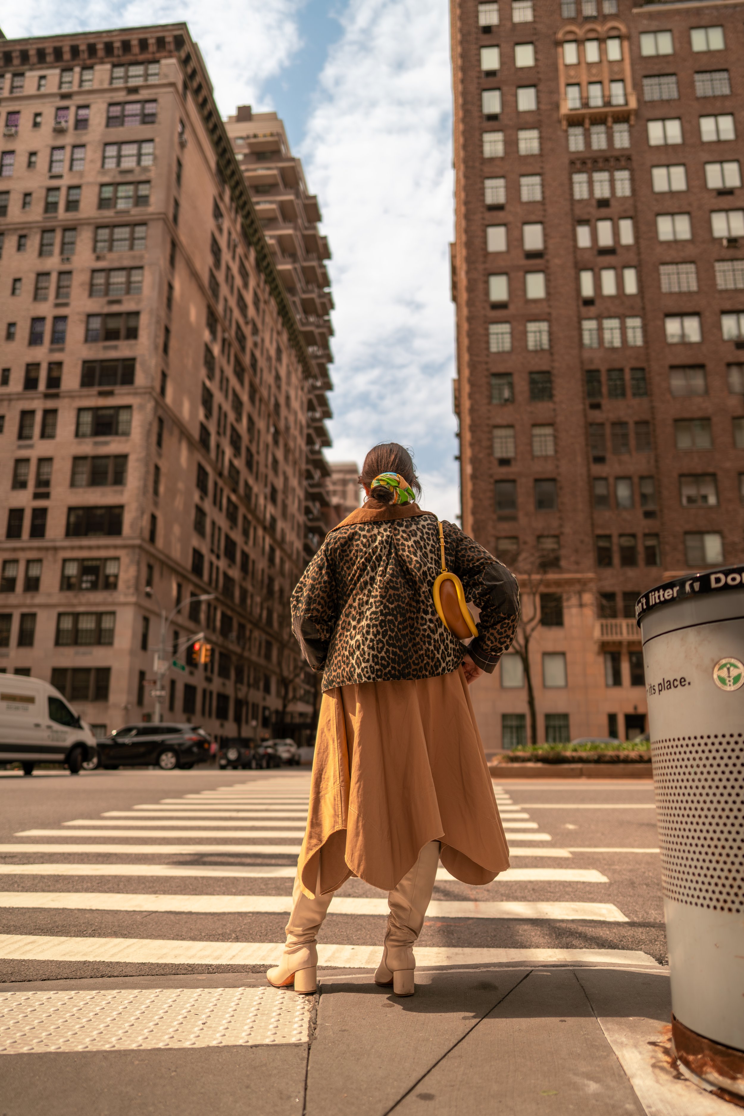 Person standing at a crosswalk in an urban city with tall buildings, wearing a leopard print jacket, a beige flowing skirt, cream-colored boots, and carrying a yellow bag shaped like a banana, with a hair bun and a green scrunchie.