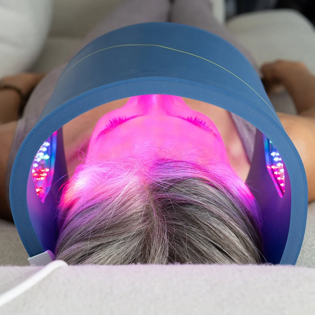 A person lying down under a pink LED light therapy device, with the camera positioned above their head showing their face with closed eyes.