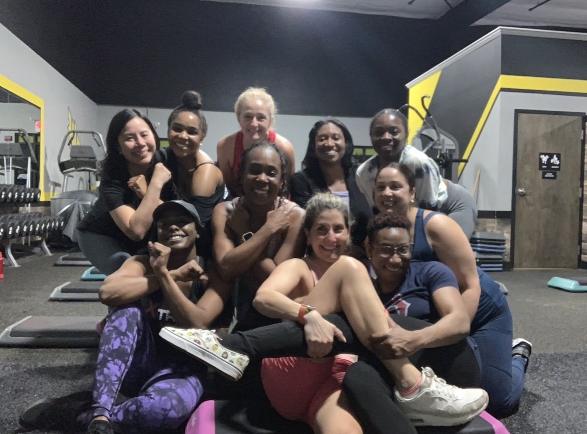 Group of women and one man posing together in a gym after a workout, smiling and showing muscle.