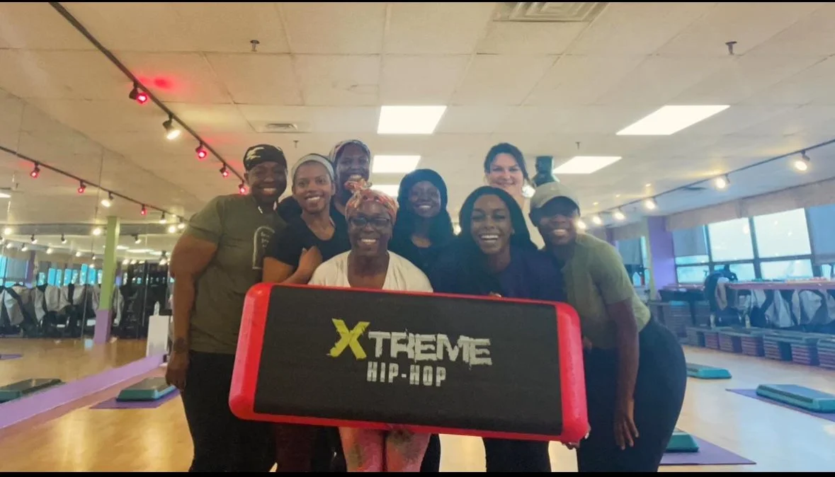 Group of smiling people at a fitness studio holding a sign that reads "Xtreme Hip-Hop" with gym equipment and exercise mats in the background.
