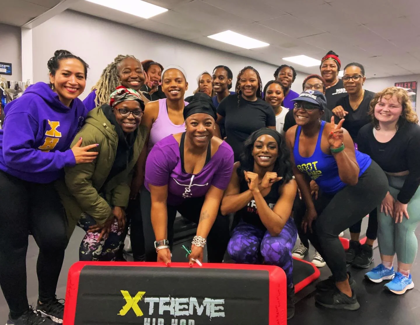 Group of women participating in a fitness class at Xtreme Hip Hop fitness gym, posing together and smiling, with some holding workout equipment.