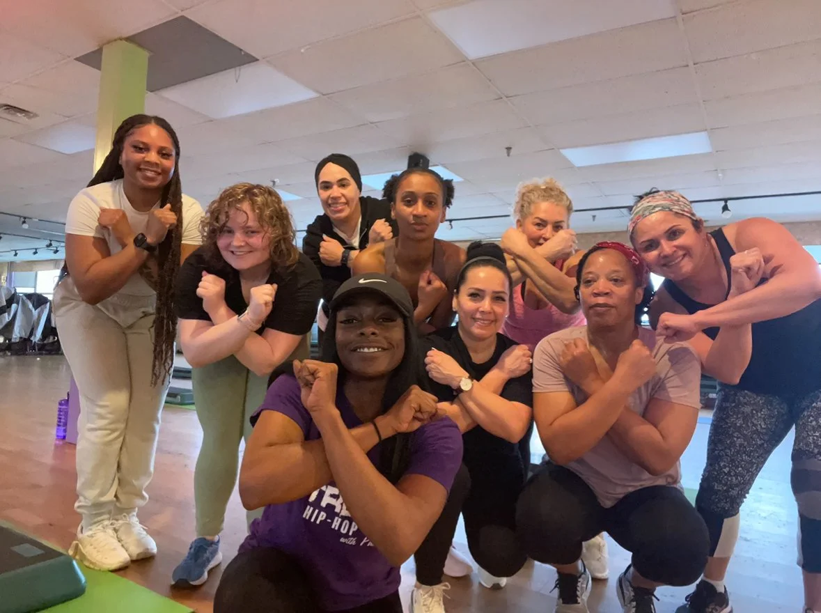 Group of nine women in fitness attire posing together in a gym, performing a strong pose with crossed arms.