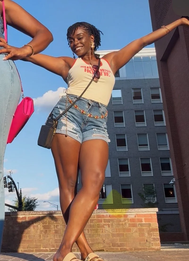 A young woman smiling with arms outstretched, wearing a tank top, denim shorts, and sunglasses hanging from her shirt in an urban outdoor setting.