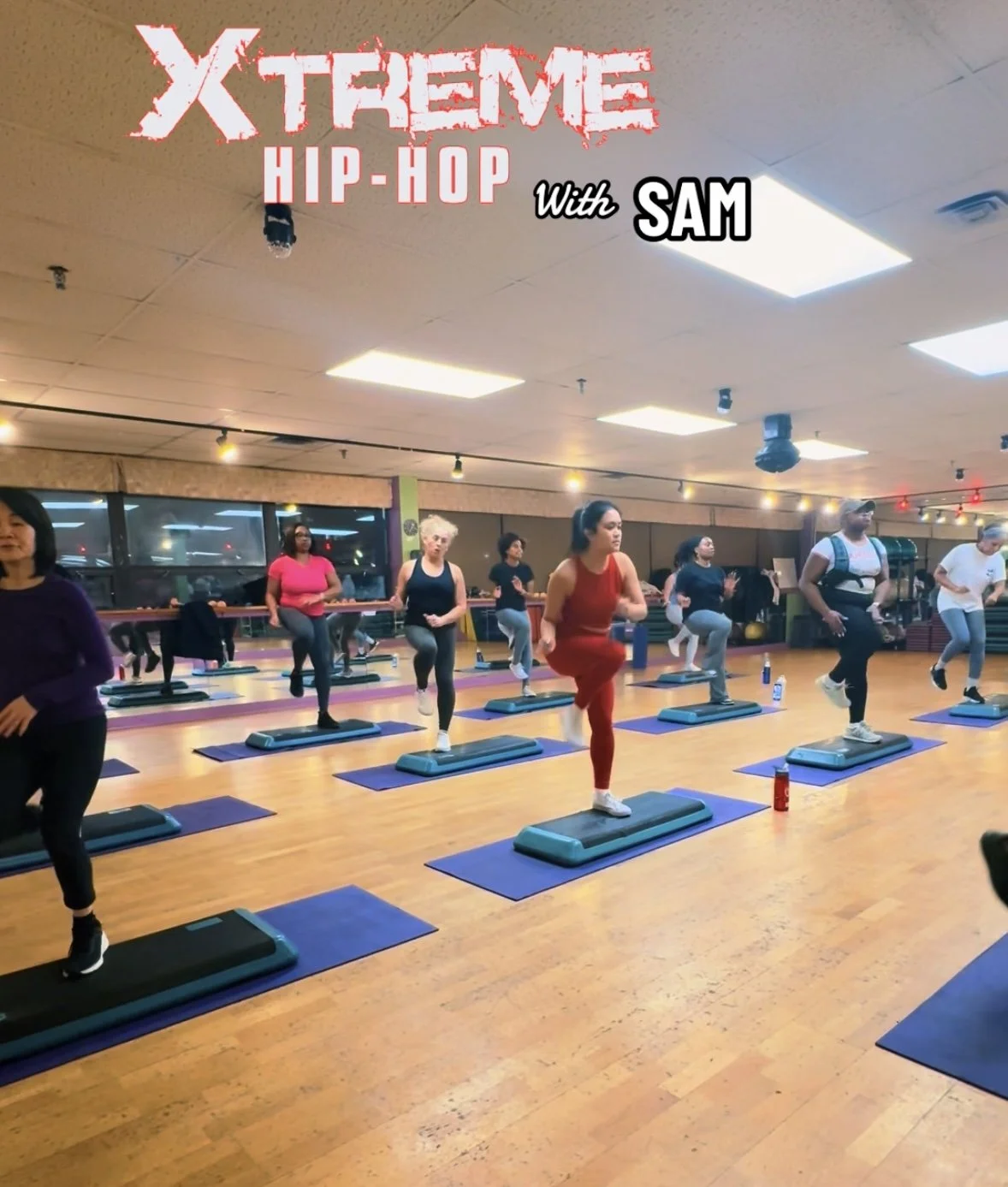 People participating in a group fitness class on step aerobics in a gym, led by an instructor, with large windows and ceiling lights.