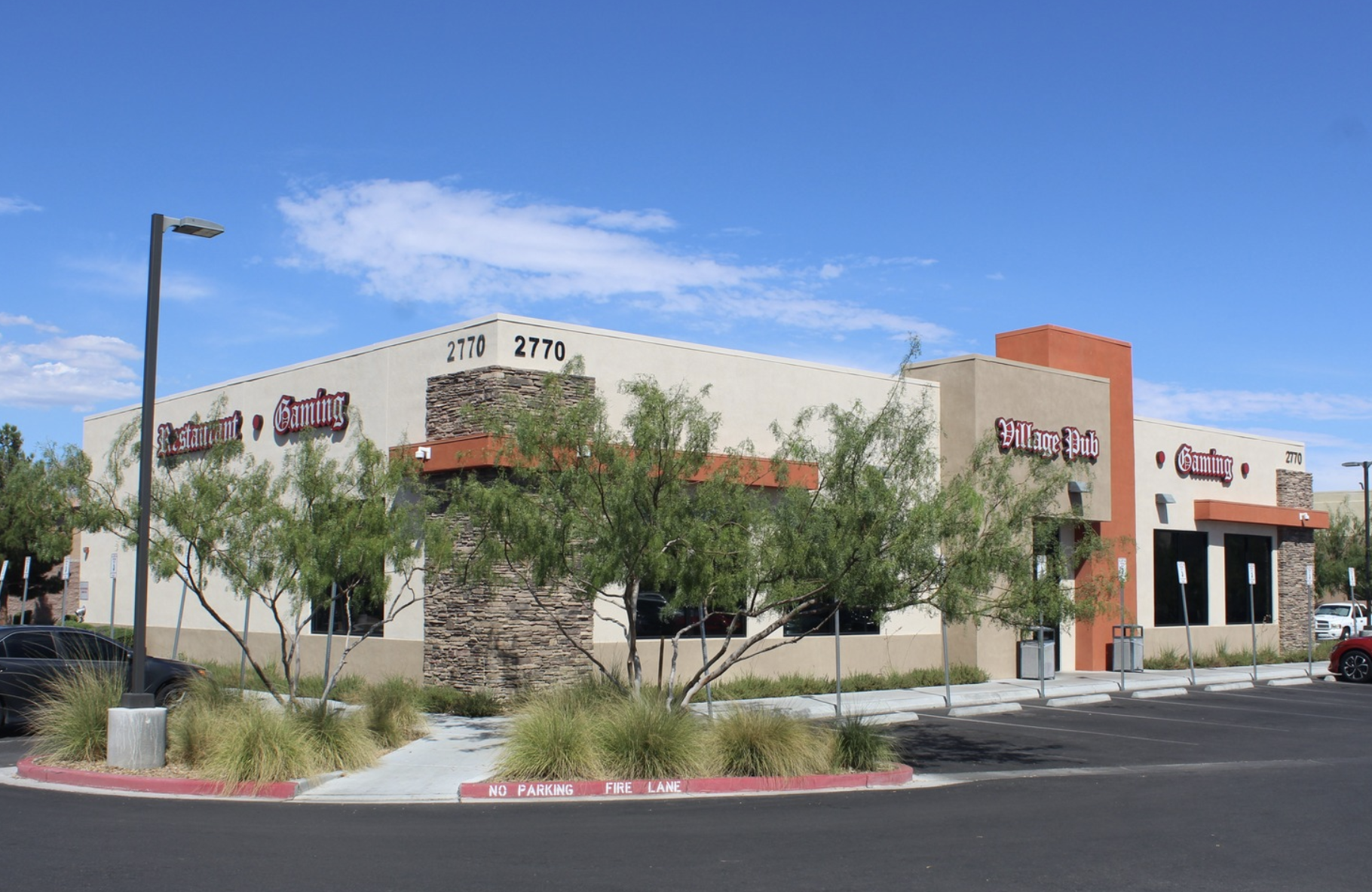 Exterior view of a modern building with signs indicating it is a restaurant, gaming, and a village pub, on a sunny day with clear blue sky and parking lot in front.