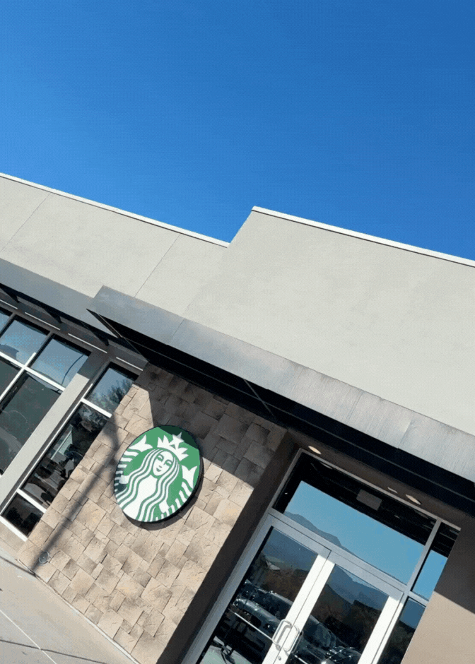 Exterior view of a Starbucks coffee shop with a large green and white logo, beige stone wall, glass door, and windows under a bright blue sky.