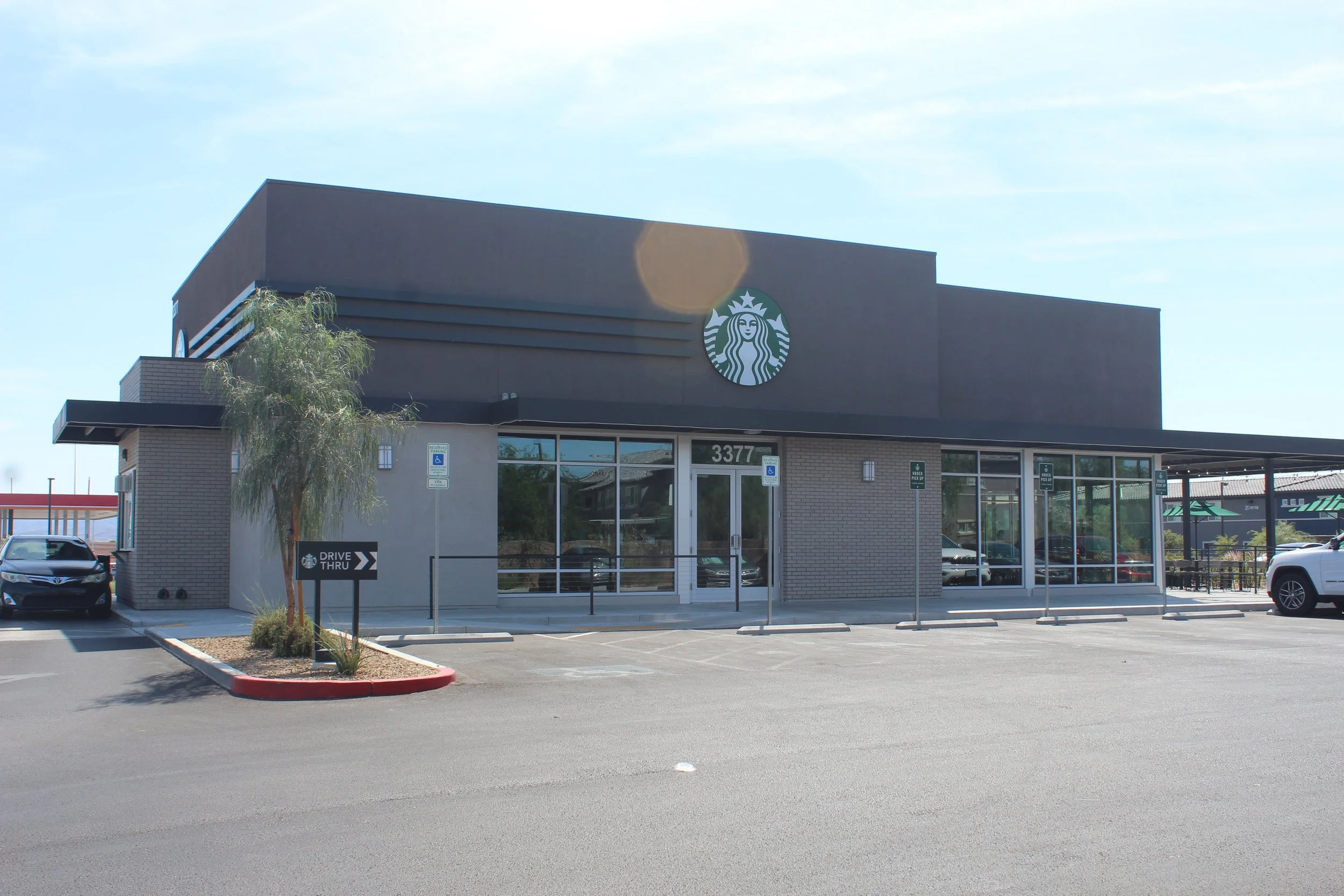 A Starbucks coffee shop with a gray exterior, large windows, and the Starbucks logo on the building. There is a small tree in front, several parking spaces, and a drive-thru sign.