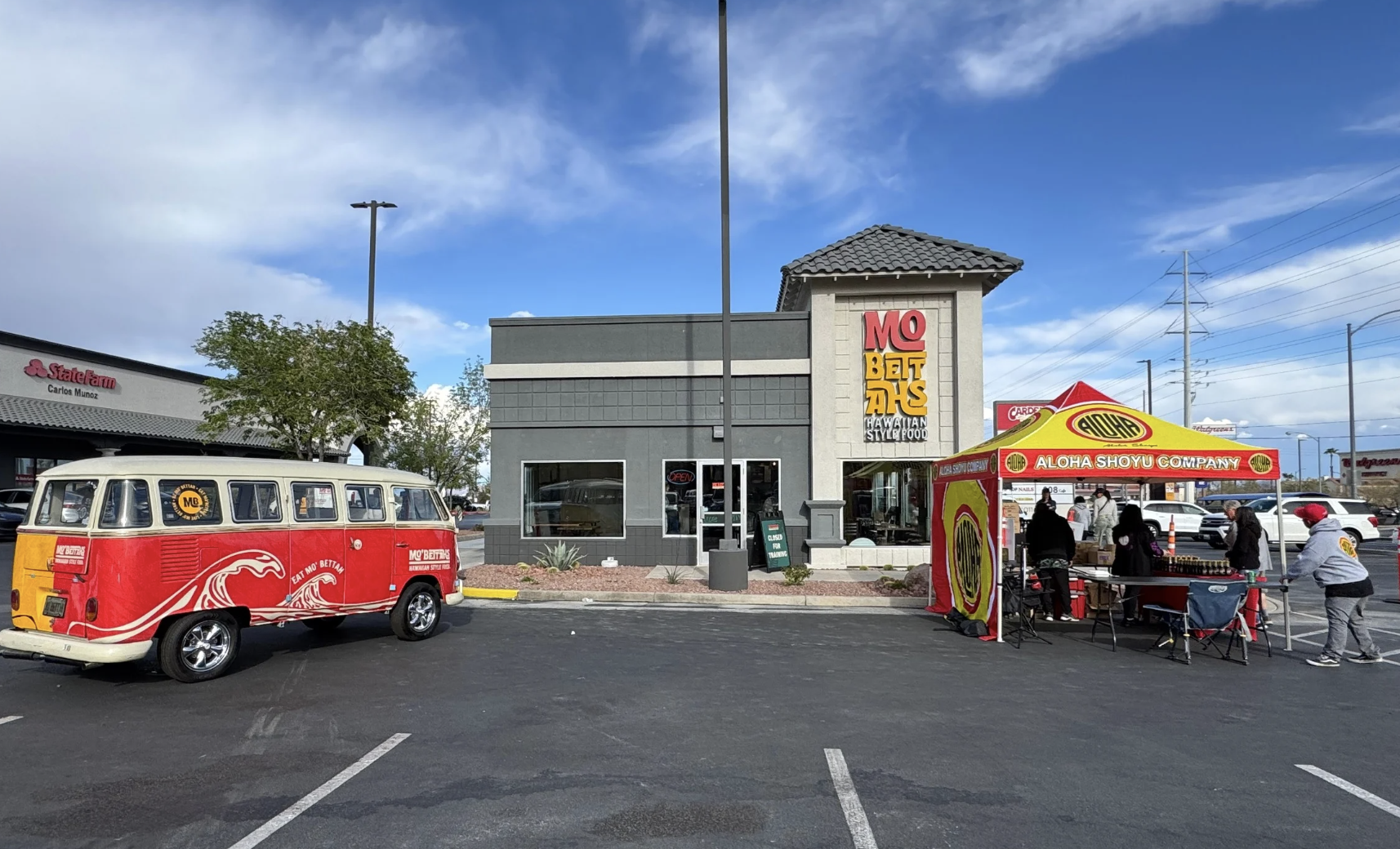 A parking lot scene in front of a restaurant with a gray building that has a sign for Hawaiian-style food, a red and yellow tent with people gathered around, a vintage red and cream van with graphics and logos, and a sign for a State Farm insurance office in the background. The sky is partly cloudy.