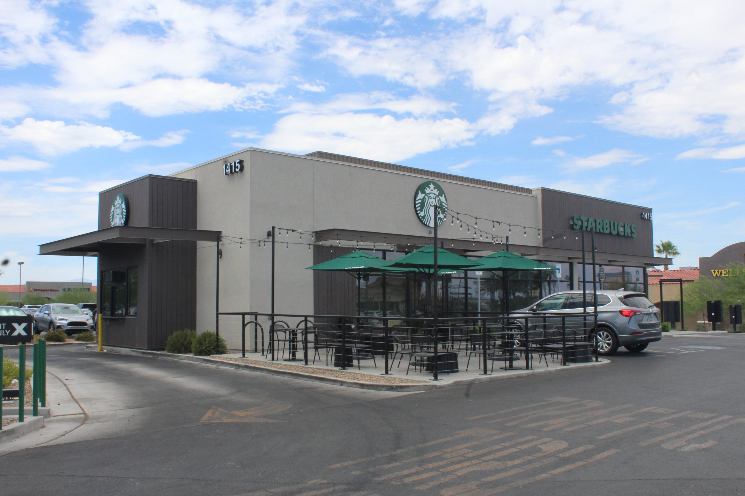 Outside view of a Starbucks coffee shop with outdoor seating under green umbrellas, parked cars, and a clear sky.