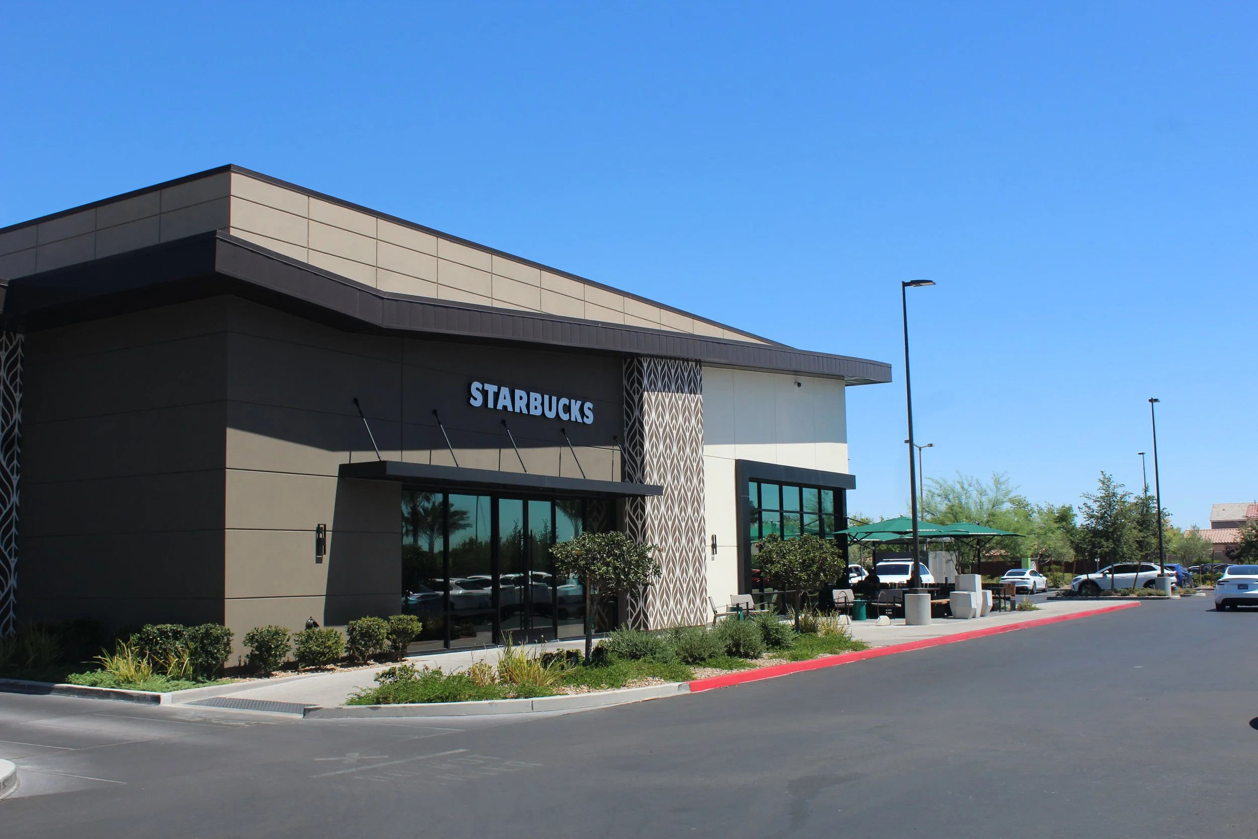 Starbucks coffee shop in a shopping center with outdoor seating, green umbrellas, and parking lot under a clear blue sky.
