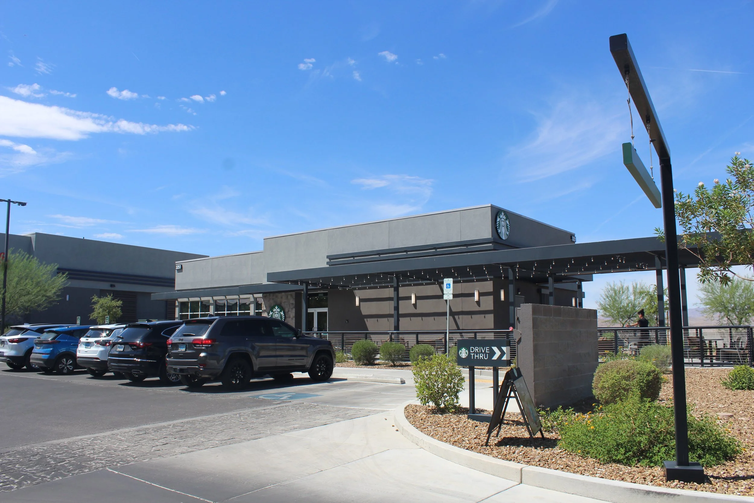A Starbucks coffee shop with a parking lot in front, some cars, and a clear blue sky. There is a drive-thru sign and a person sitting on an outdoor patio.