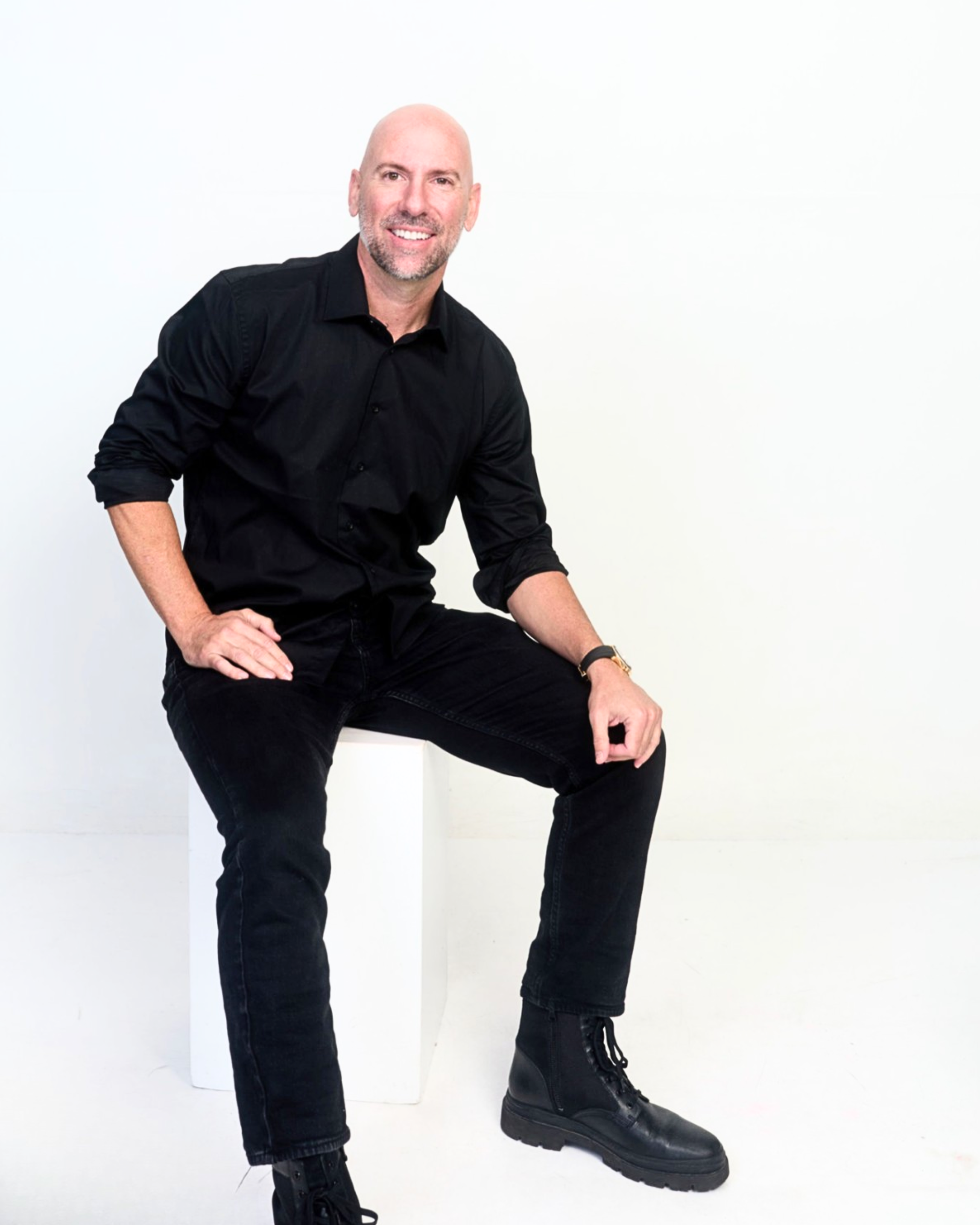 A smiling bald man with a beard wearing a black shirt, black pants, and black combat boots, sitting on a white cube against a plain white background.