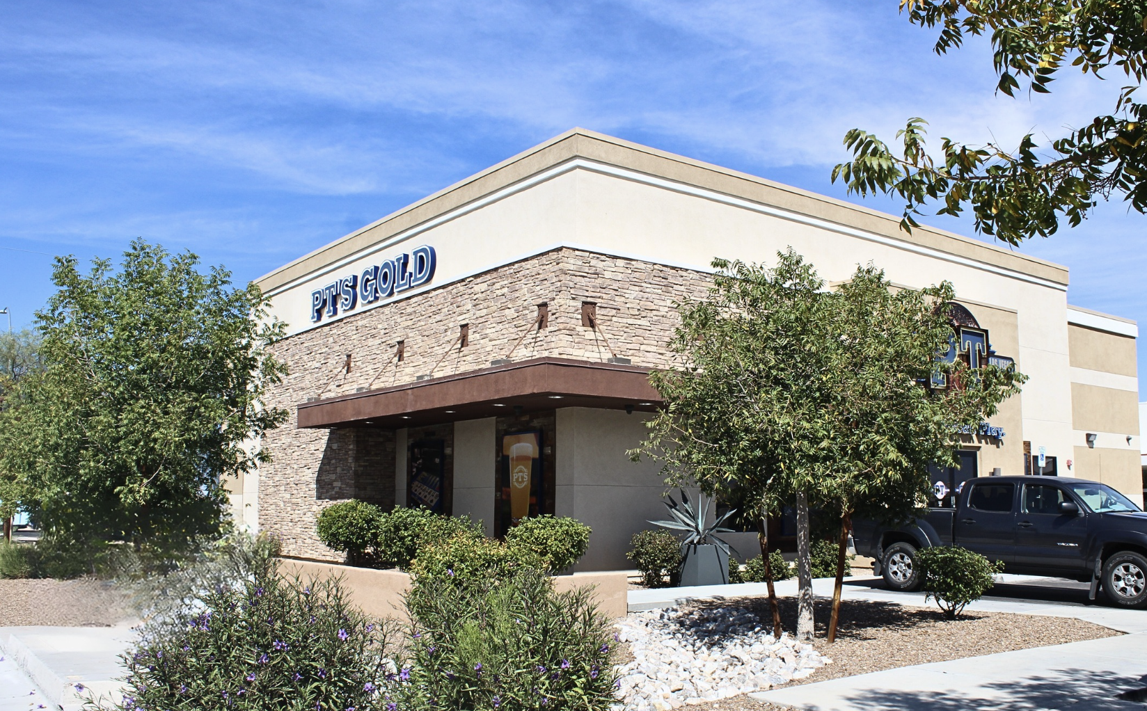 Exterior view of a PTS Gold store with a beige building, stone accents, trees, shrubs, and a parked black pickup truck under a blue sky.