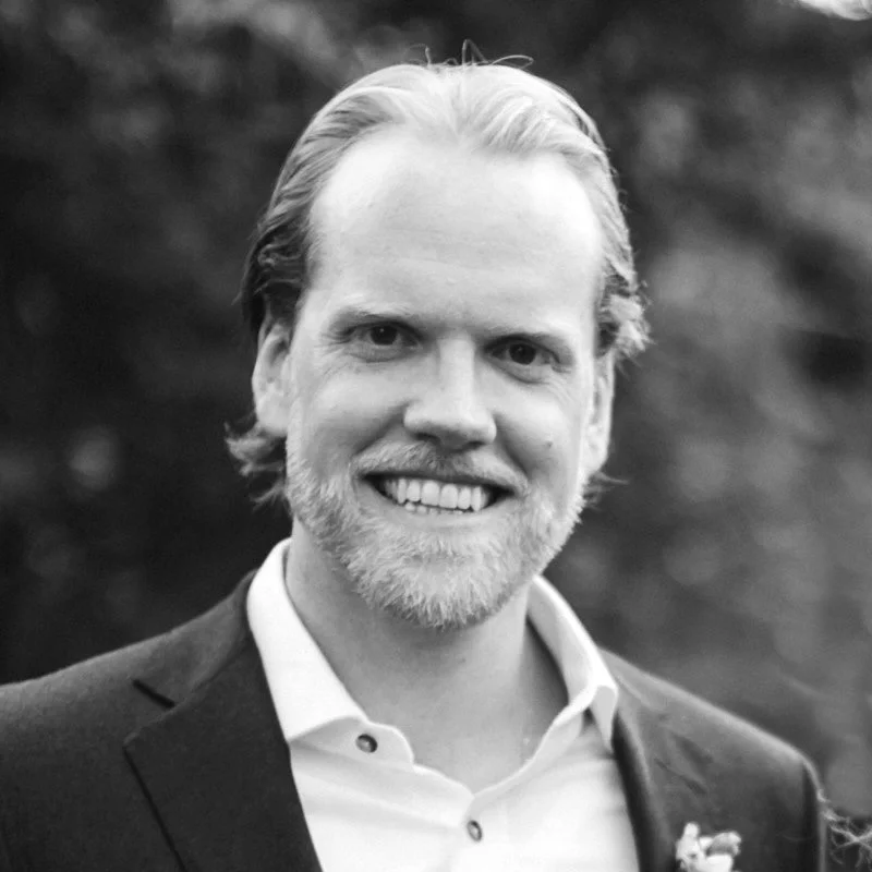 A smiling man with light hair and a beard, wearing a suit jacket and a white shirt, outdoors with a blurred natural background.