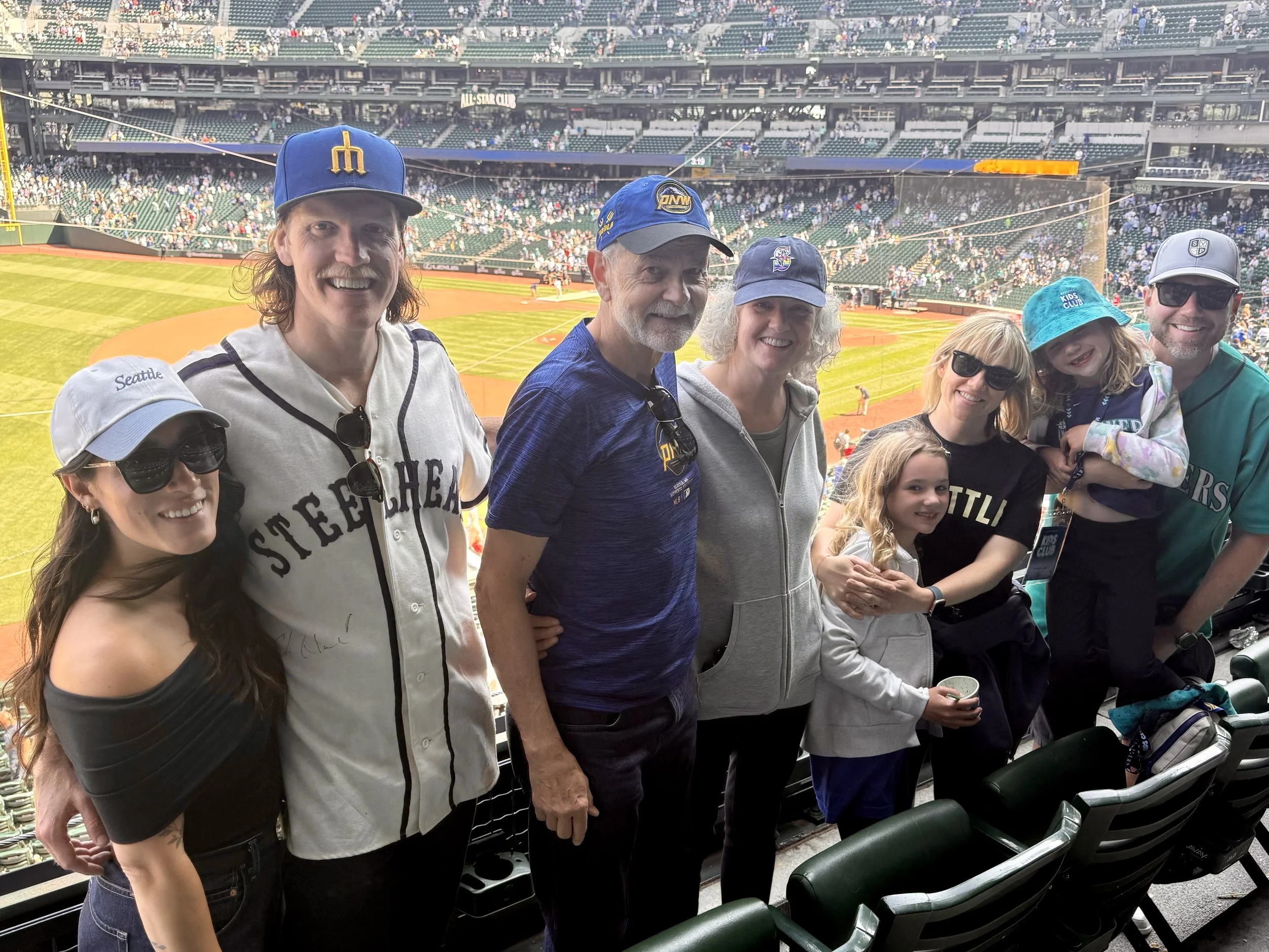Group of people at a baseball game in a stadium, some wearing Seattle Mariners gear, standing in the stands with the baseball field in the background.