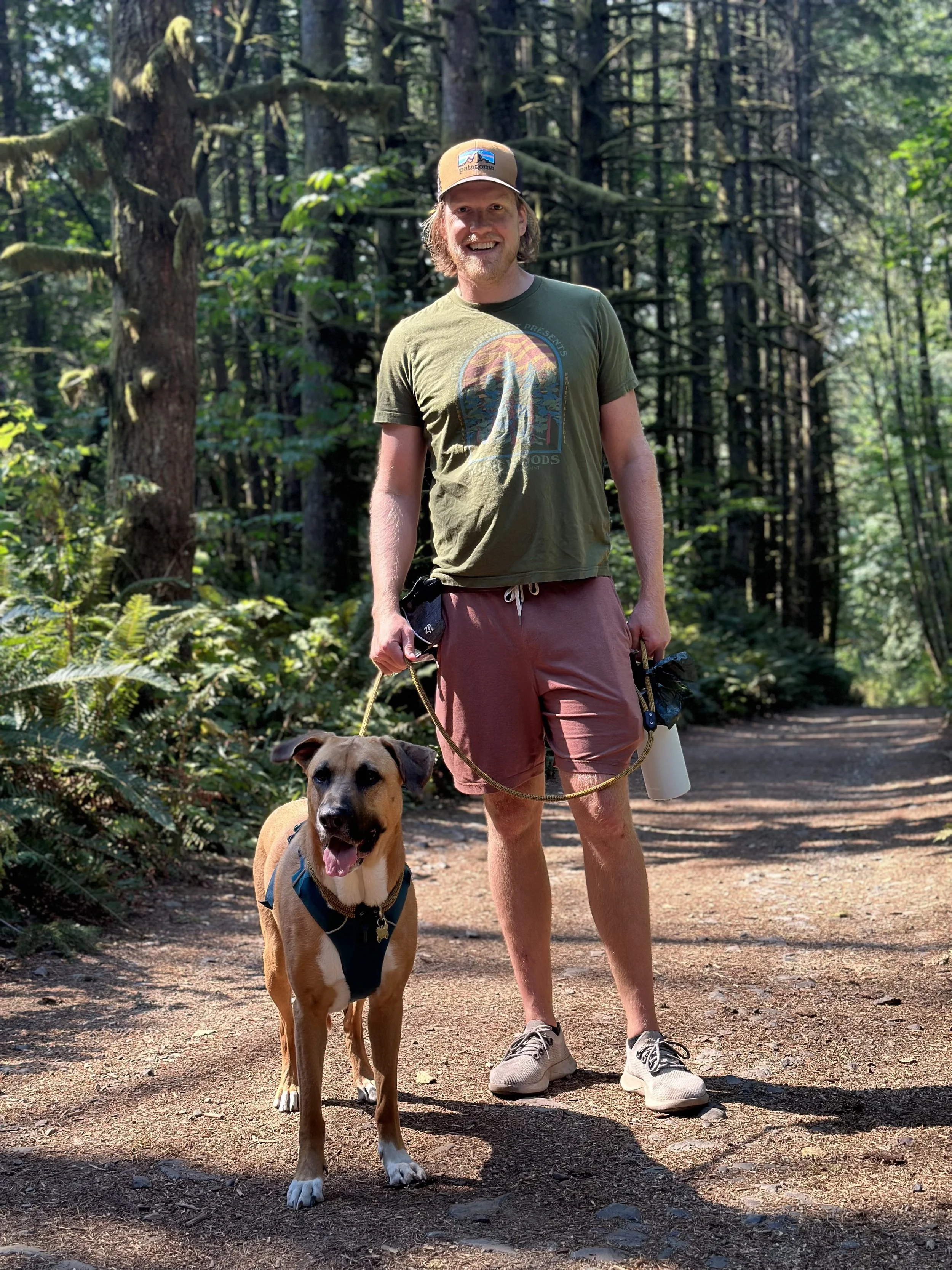 A man in a green T-shirt and pink shorts walking a brown and white dog on a trail in a forest.