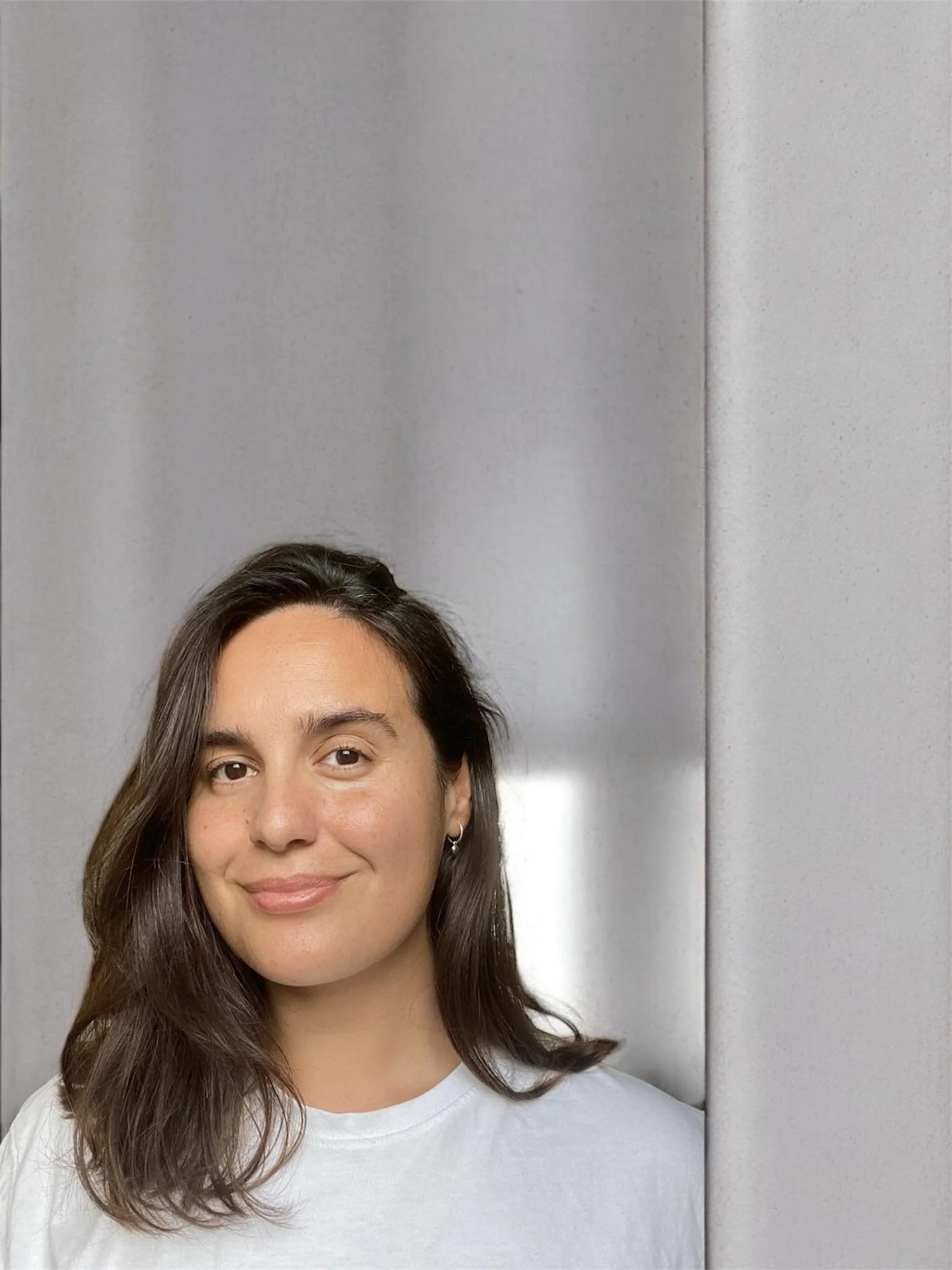 A woman with shoulder-length brown hair, wearing a white shirt, smiling softly, standing in front of a light gray wall.