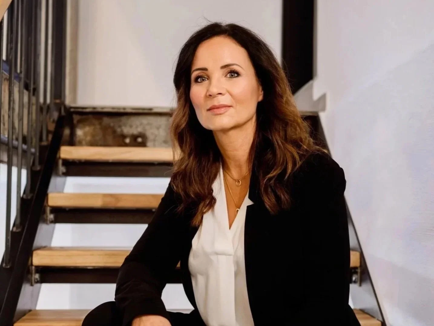 Eine Frau sitzt auf einer Holztreppe, trägt einen schwarzen Blazer und eine weiße Bluse, hat lange braune Haare und schaut in die Kamera.