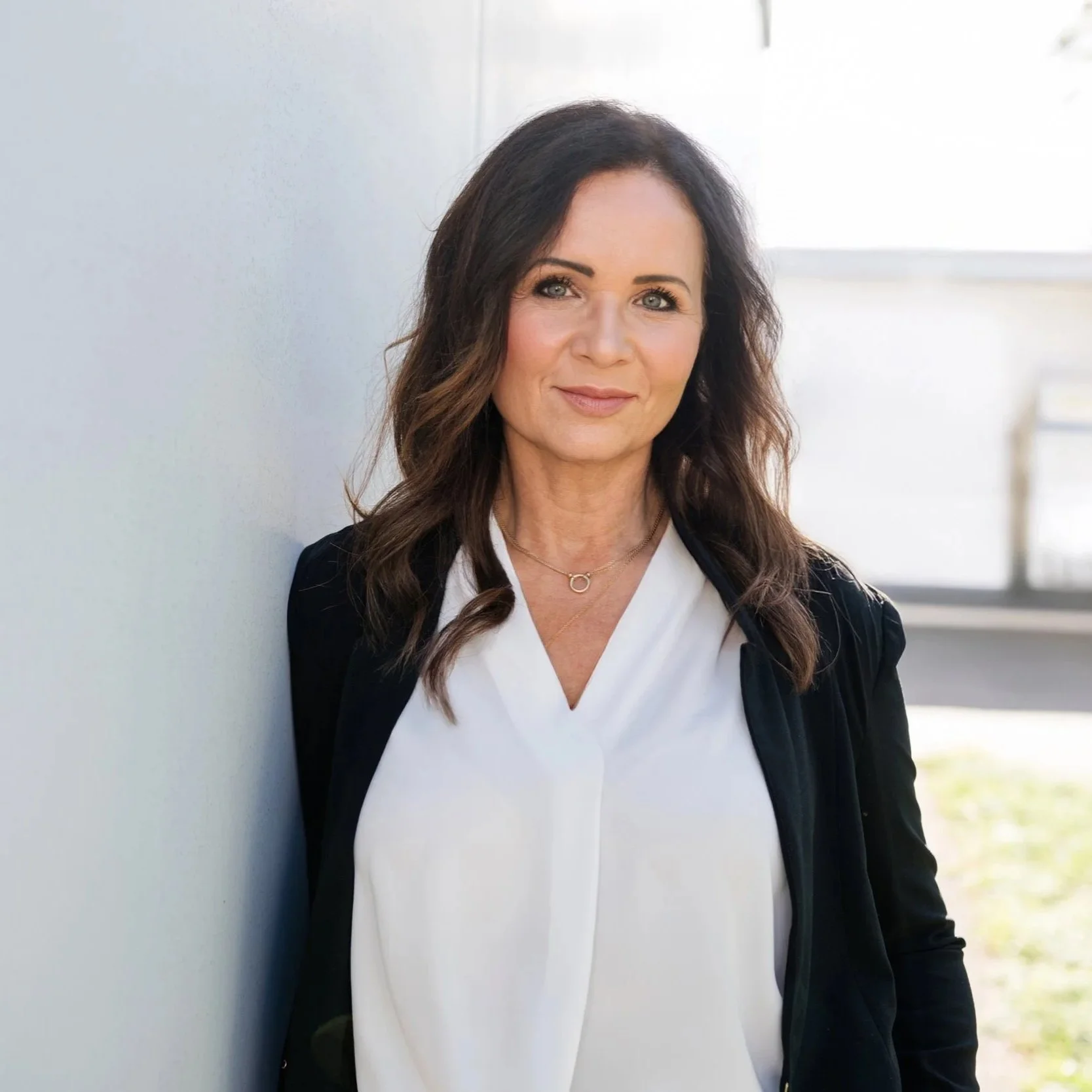 Eine professionell wirkende Frau mit braunen, lockigen Haaren steht an einer weißen Wand im Freien, trägt ein weißes Hemd und eine schwarze Jacke, lächelt leicht.