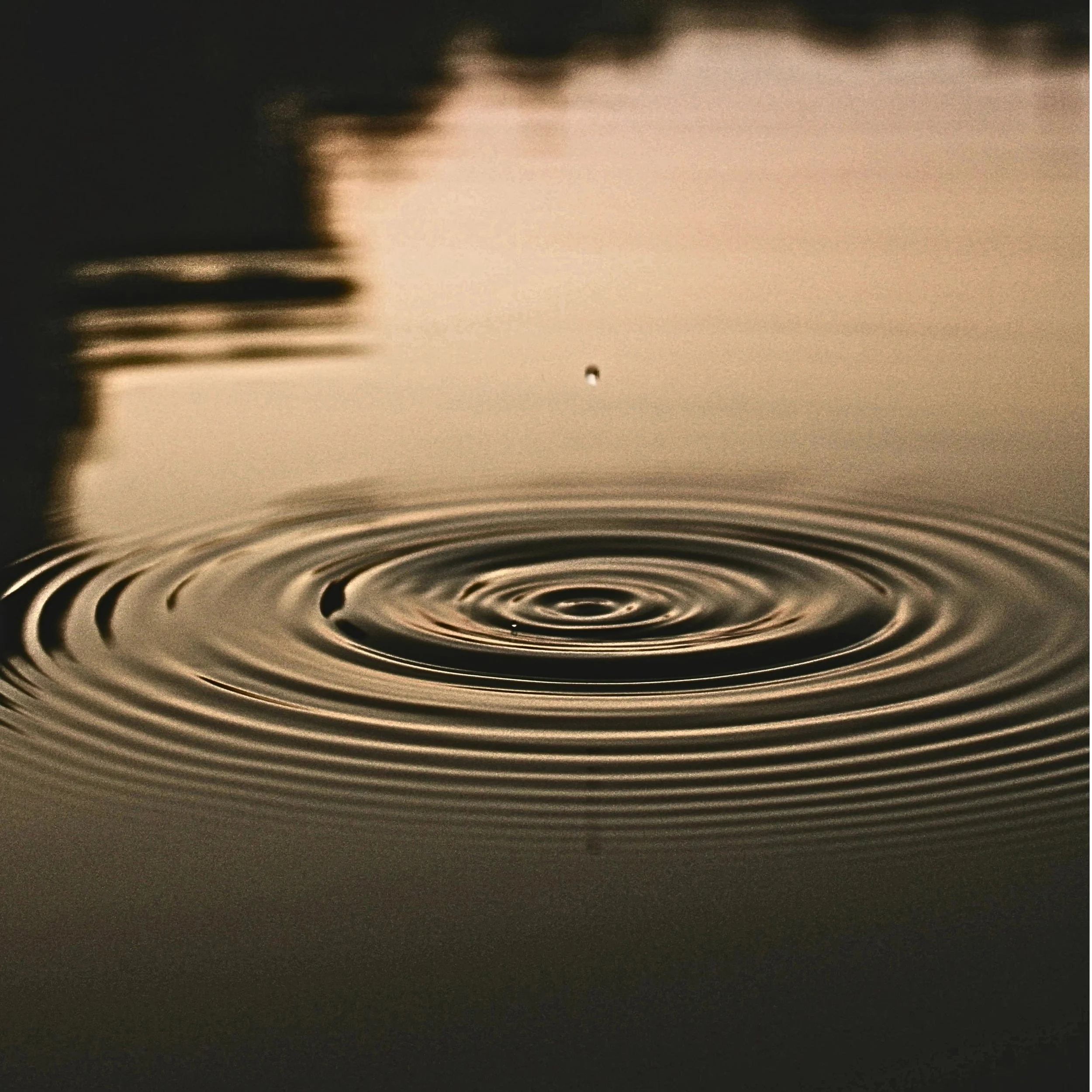 Ein Wassertropfen fällt in eine Wasseroberfläche und verursacht kreisförmige Wellen. Die Szene steht für innere Balance und seelischen Tiefgang.