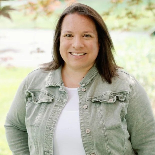 A woman with shoulder-length dark brown hair, smiling, wearing a light gray denim jacket over a white top, standing outdoors with greenery in the background.
