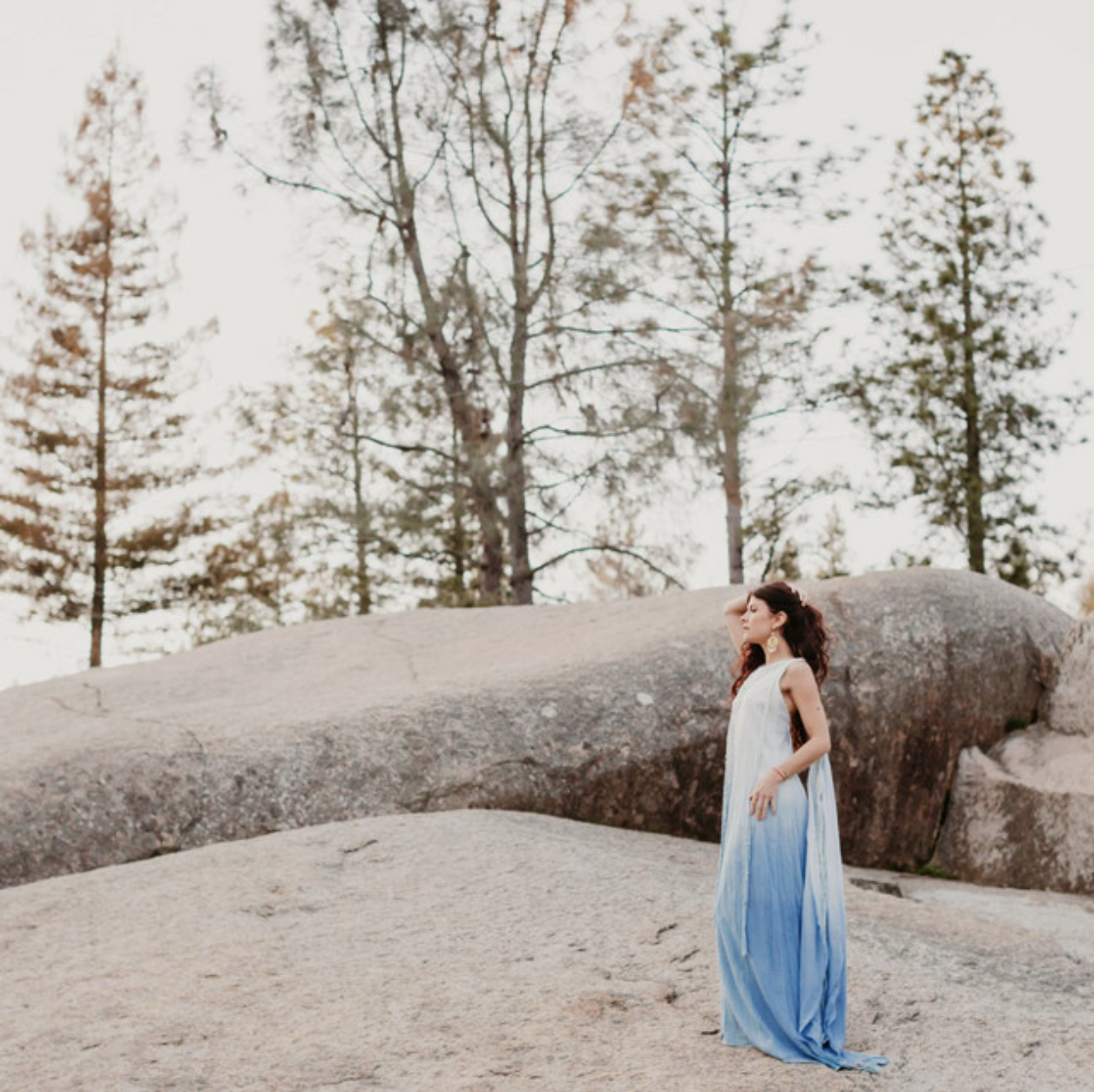 A woman in a flowing white and blue dress standing on rocky terrain with trees in the background, looking to the side with her hand on her face.