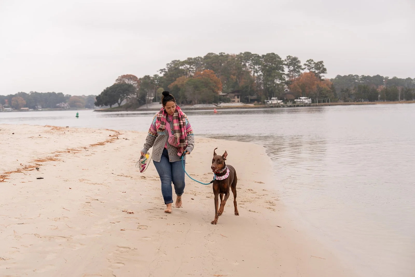 A woman walking a brown dog on a sandy beach by a calm water body during overcast weather.