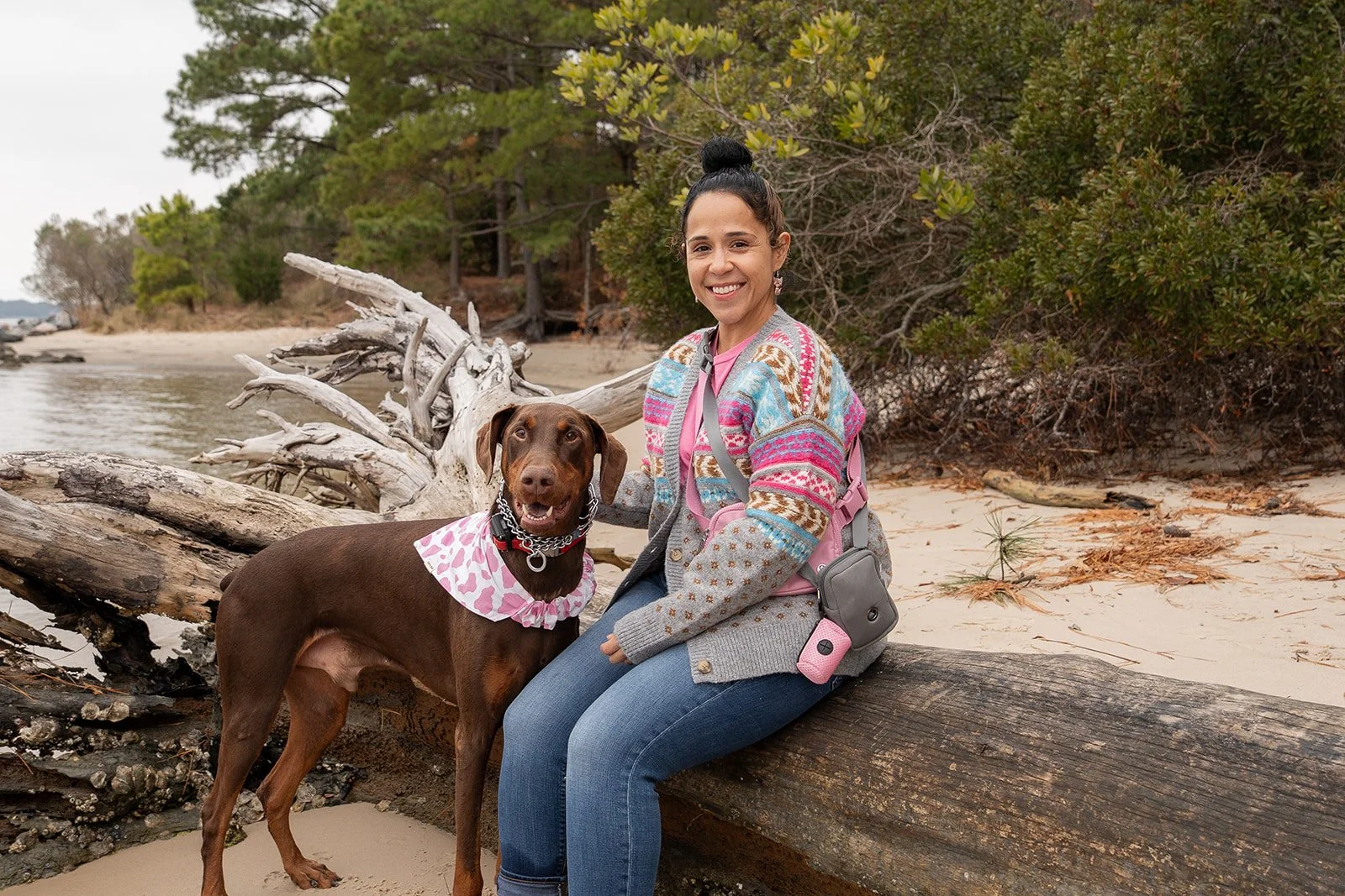 A woman sitting on a large log on a beach with a brown dog. Both are looking happy, with the woman smiling at the camera. The woman has dark hair tied up, and is wearing a colorful patterned sweater, pink shirt, and jeans. The dog is wearing a pink polka-dot bandana and a black collar. In the background, there's a shoreline with trees and branches, and driftwood on the sand.
