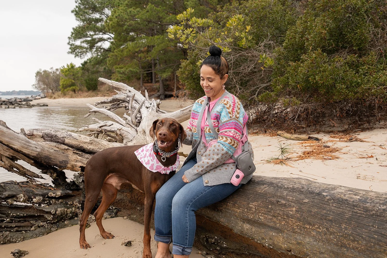 A woman in a colorful sweater sitting on a fallen log on a beach, smiling at her brown dog with a pink bandana, nearby driftwood, sand, trees, and water in the background.