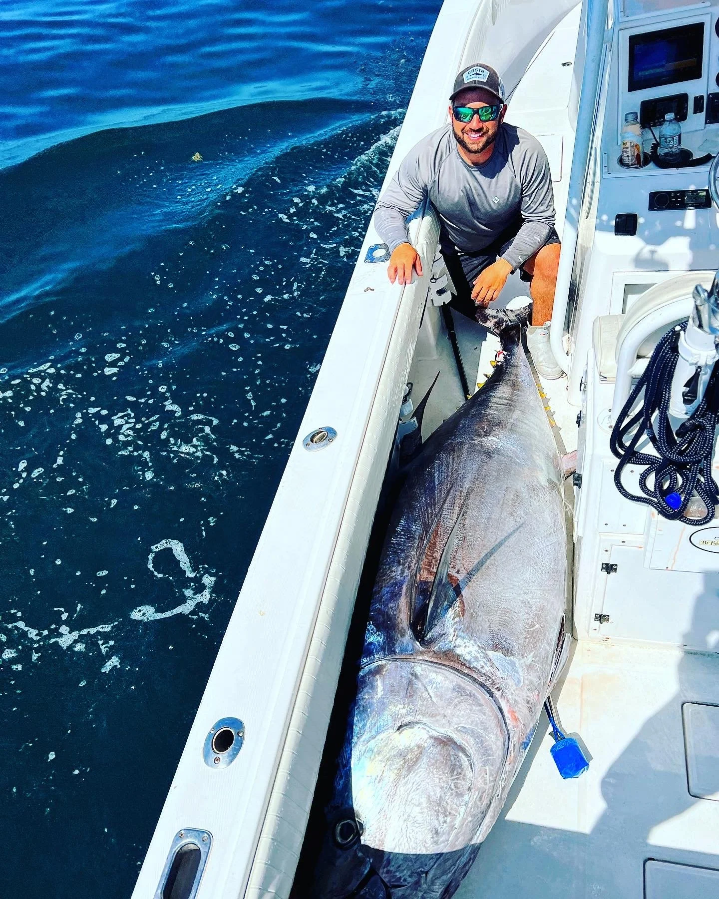 A man wearing sunglasses, a gray shirt, and dark shorts smiling on a boat with a large fish, likely a tuna, lying beside him in the boat.