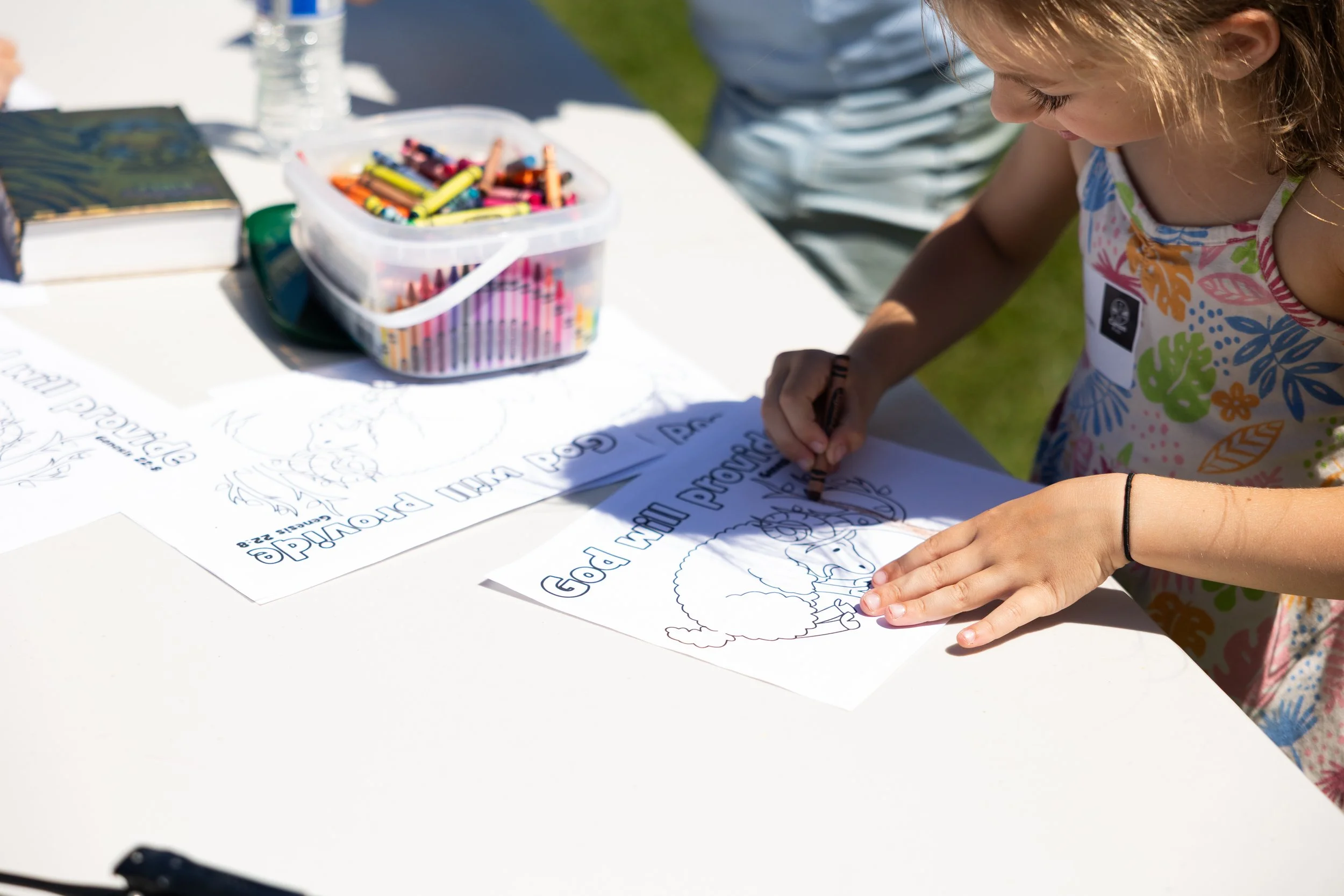 A young girl in a colorful dress is coloring a turkey outline on a piece of paper at an outdoor event. There is a plastic container with crayons and some flyers on the table.