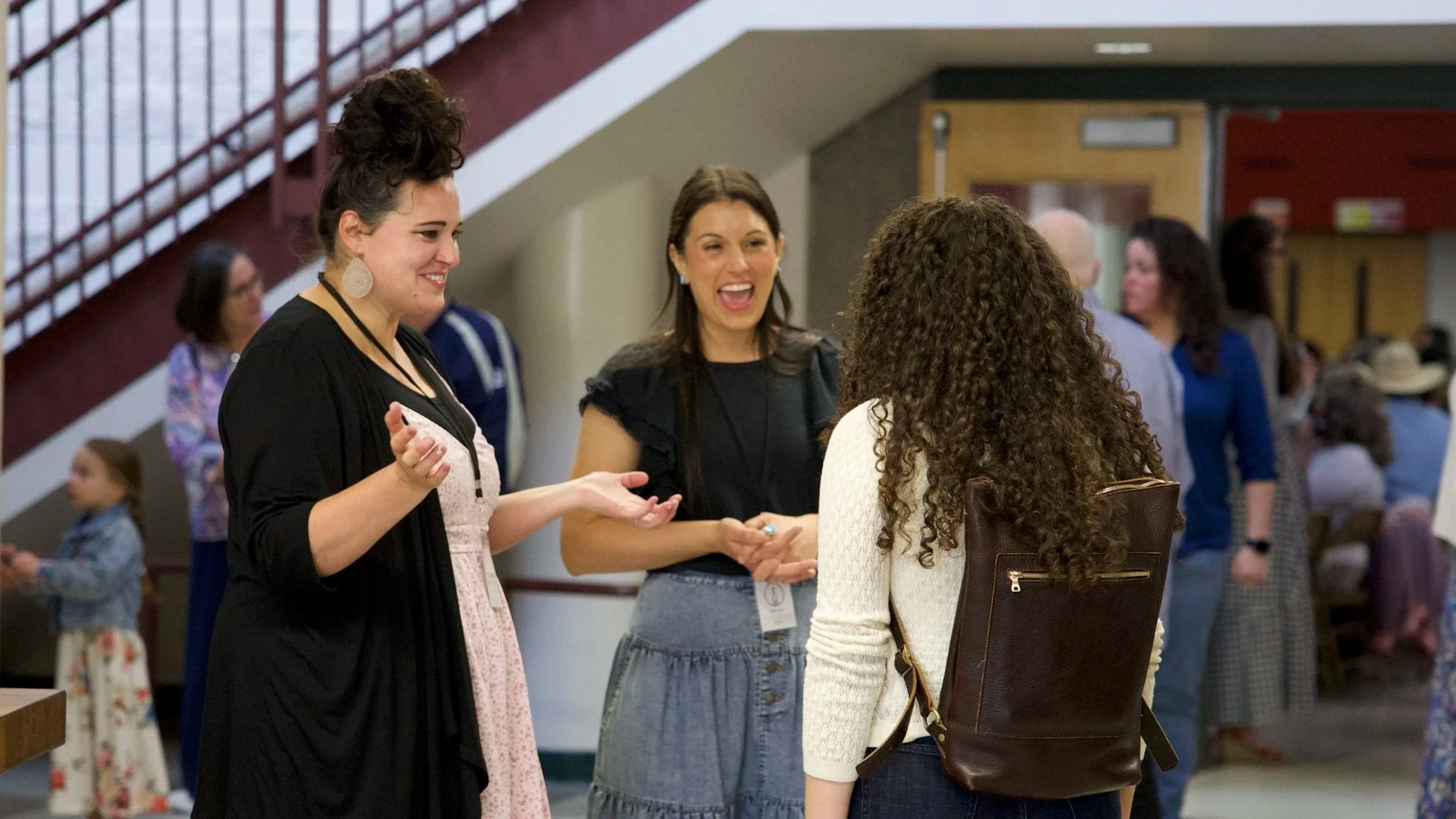 Four women engaged in conversation at an indoor event, with a background of other attendees, including children and adults, near stairs and walls.