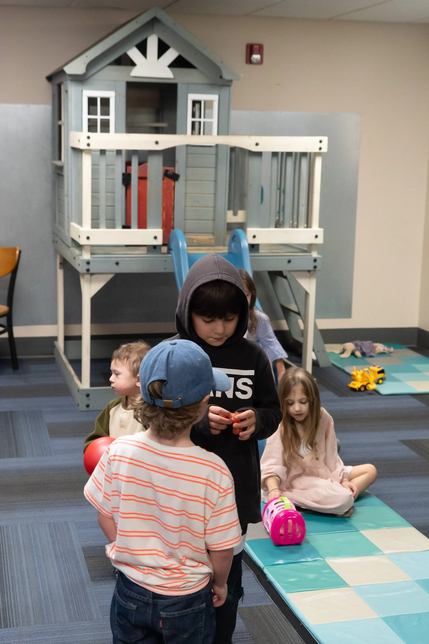 Children playing in an indoor play area with a small blue playhouse, slide, and mats.