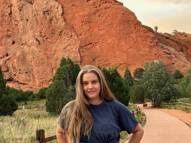 A young woman with long brown hair wearing a navy blue t-shirt stands on a pathway in a scenic desert landscape with red rock formations and green trees.