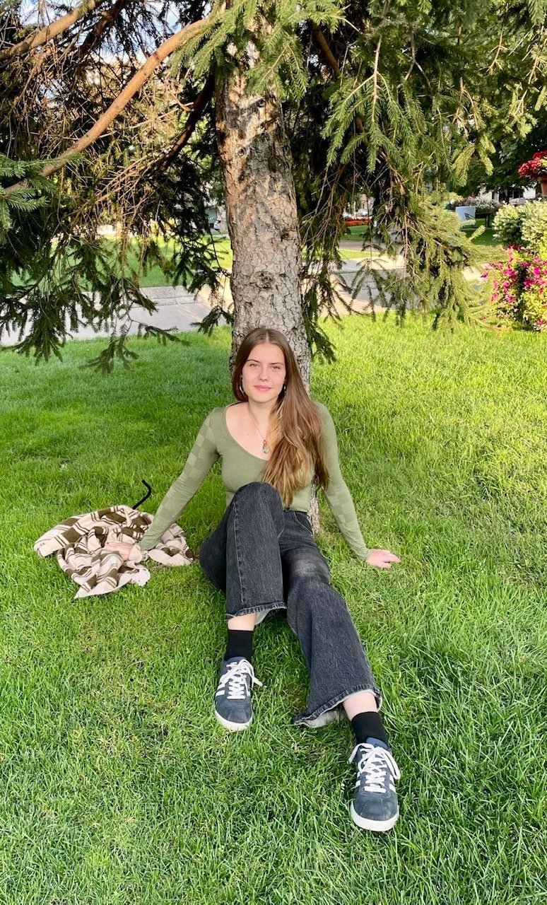 Young woman sitting on green grass with back against a large tree, wearing a green long sleeve shirt, black jeans, and sneakers, outdoors in a park or garden setting.