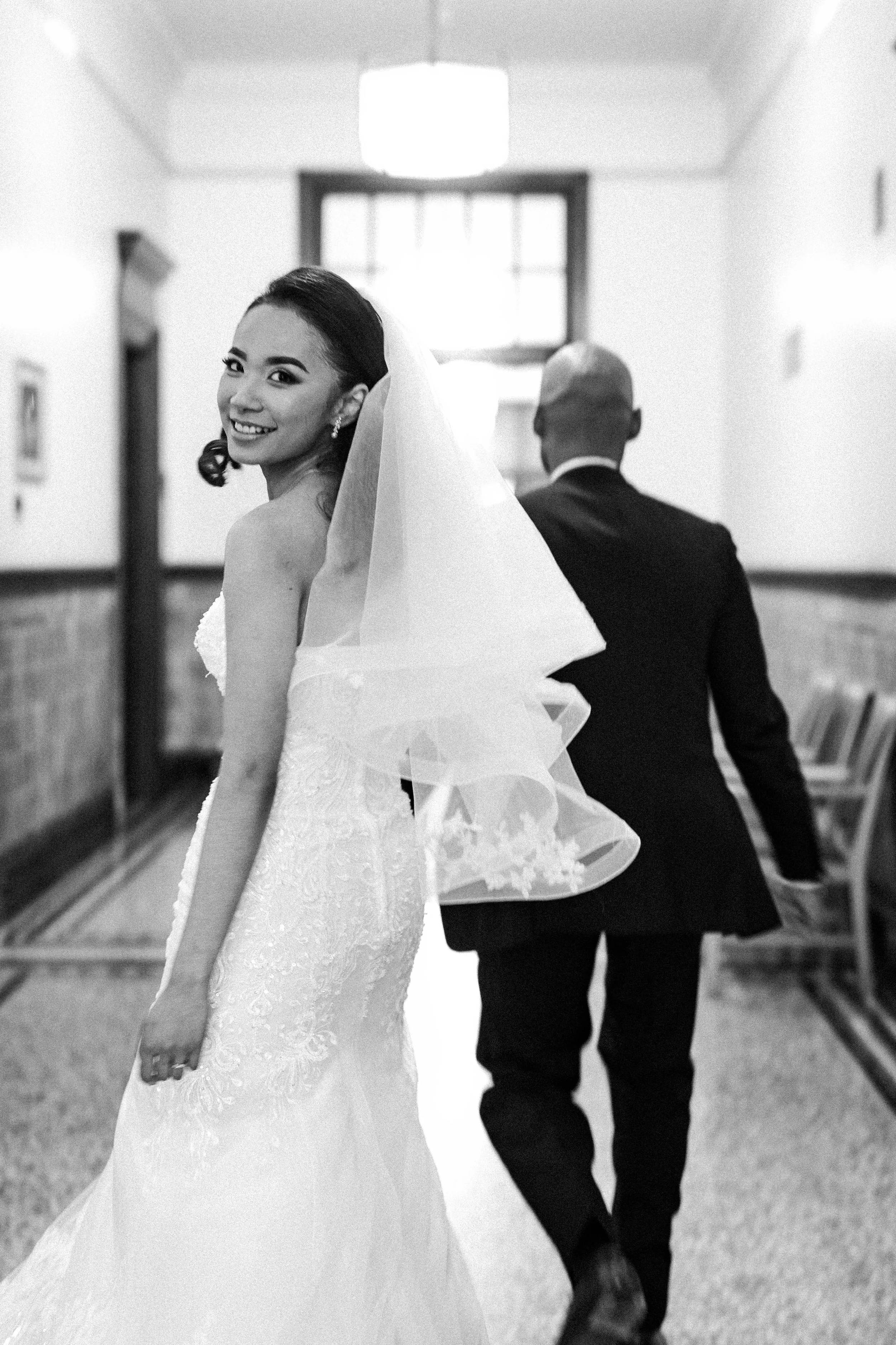 A bride in a wedding dress smiling and looking over her shoulder, with a veil, and a man in a suit walking away in a hallway.