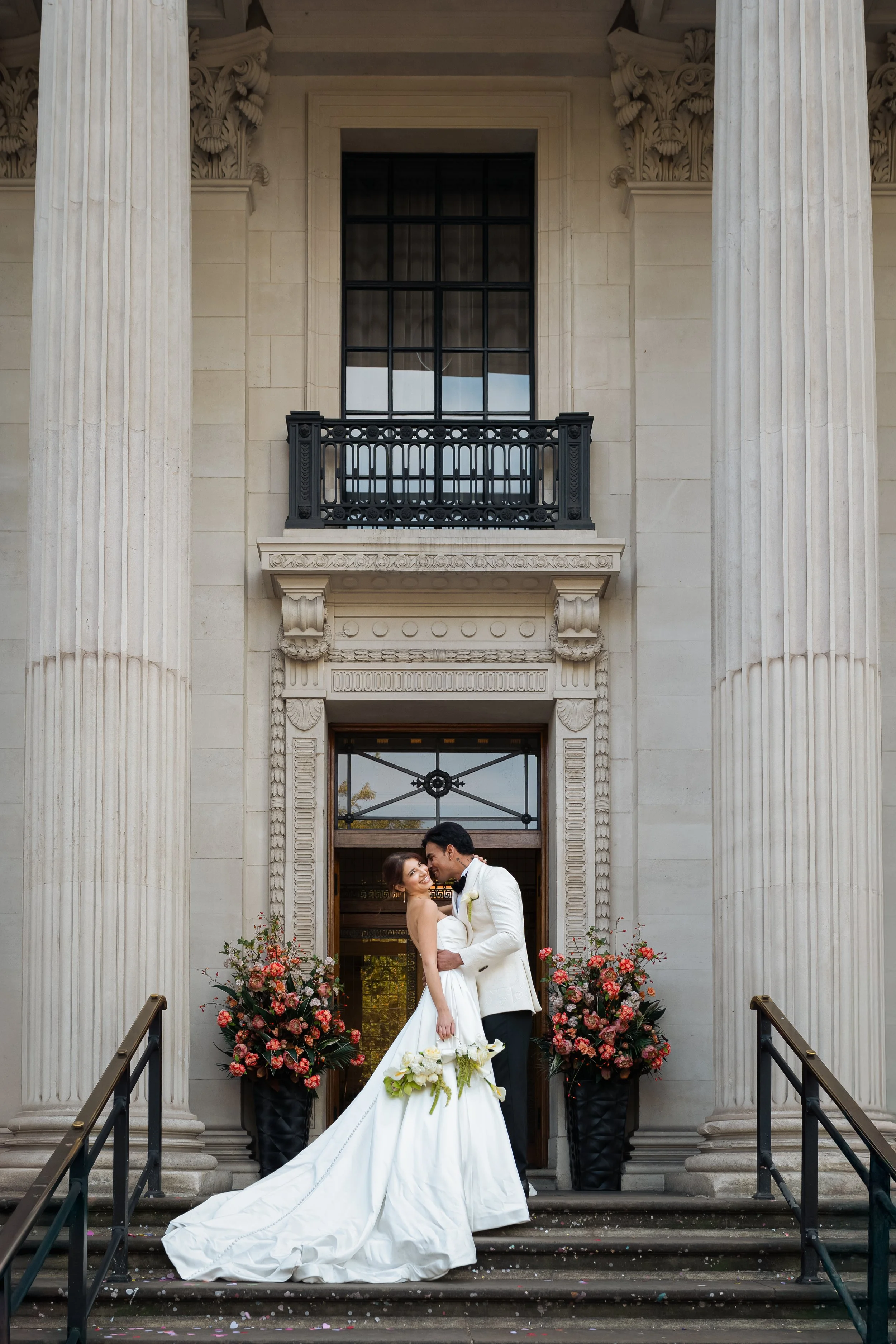 A bride and groom kissing on the steps of a building with large columns and decorated with flower arrangements.