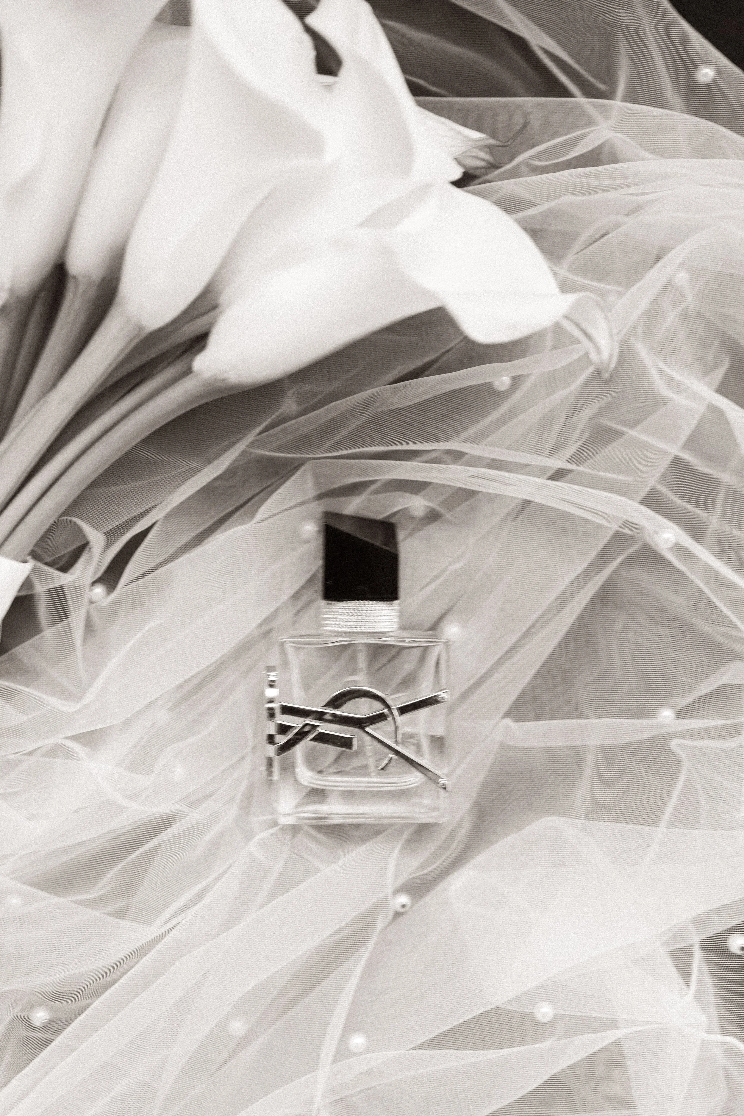 A small bottle of Chanel perfume with a gold-colored ring and chain accessory, surrounded by sheer white fabric and flowers, likely calla lilies.