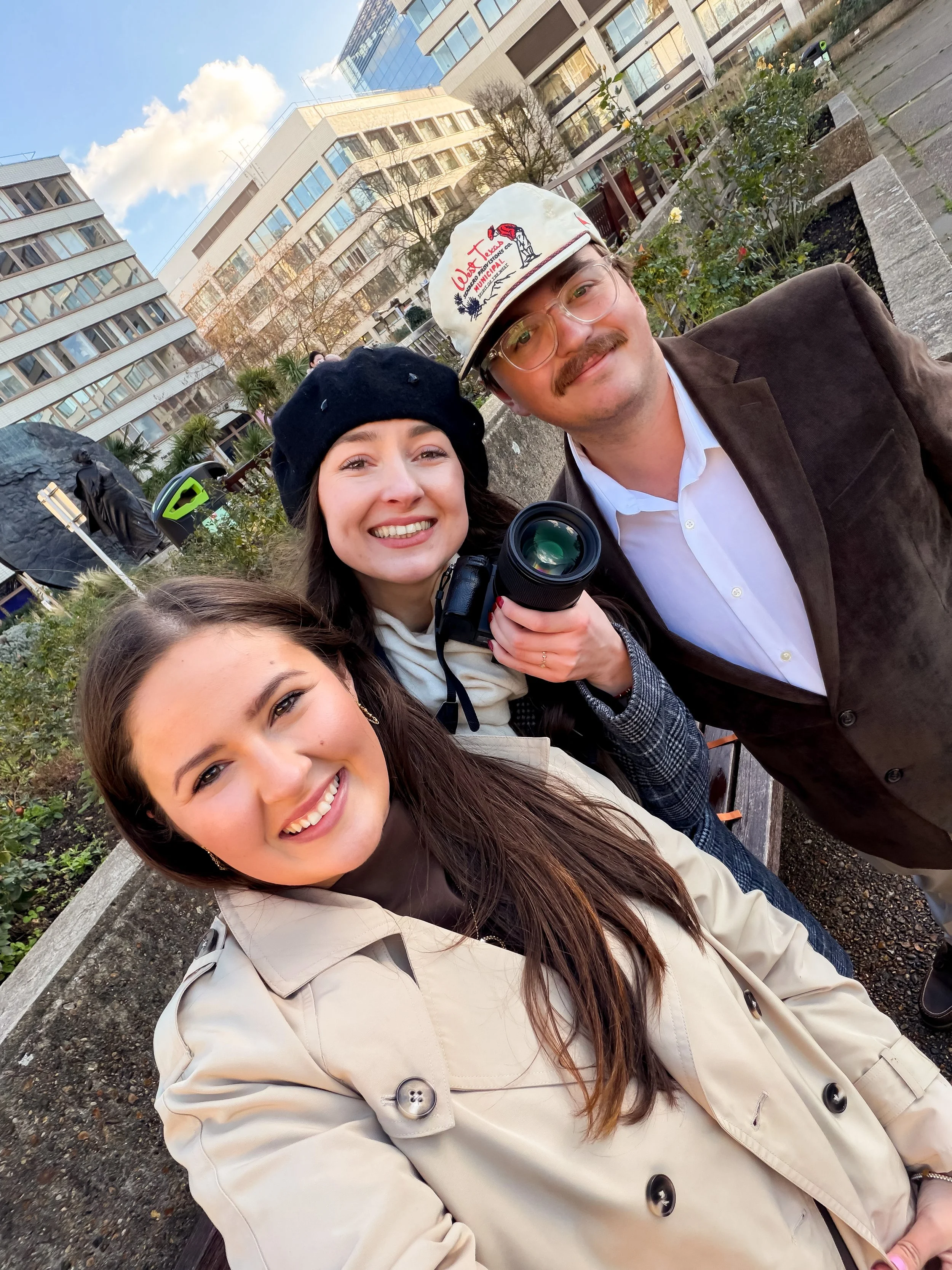 Three people taking a selfie outdoors in an urban park area with buildings and greenery, smiling at the camera.