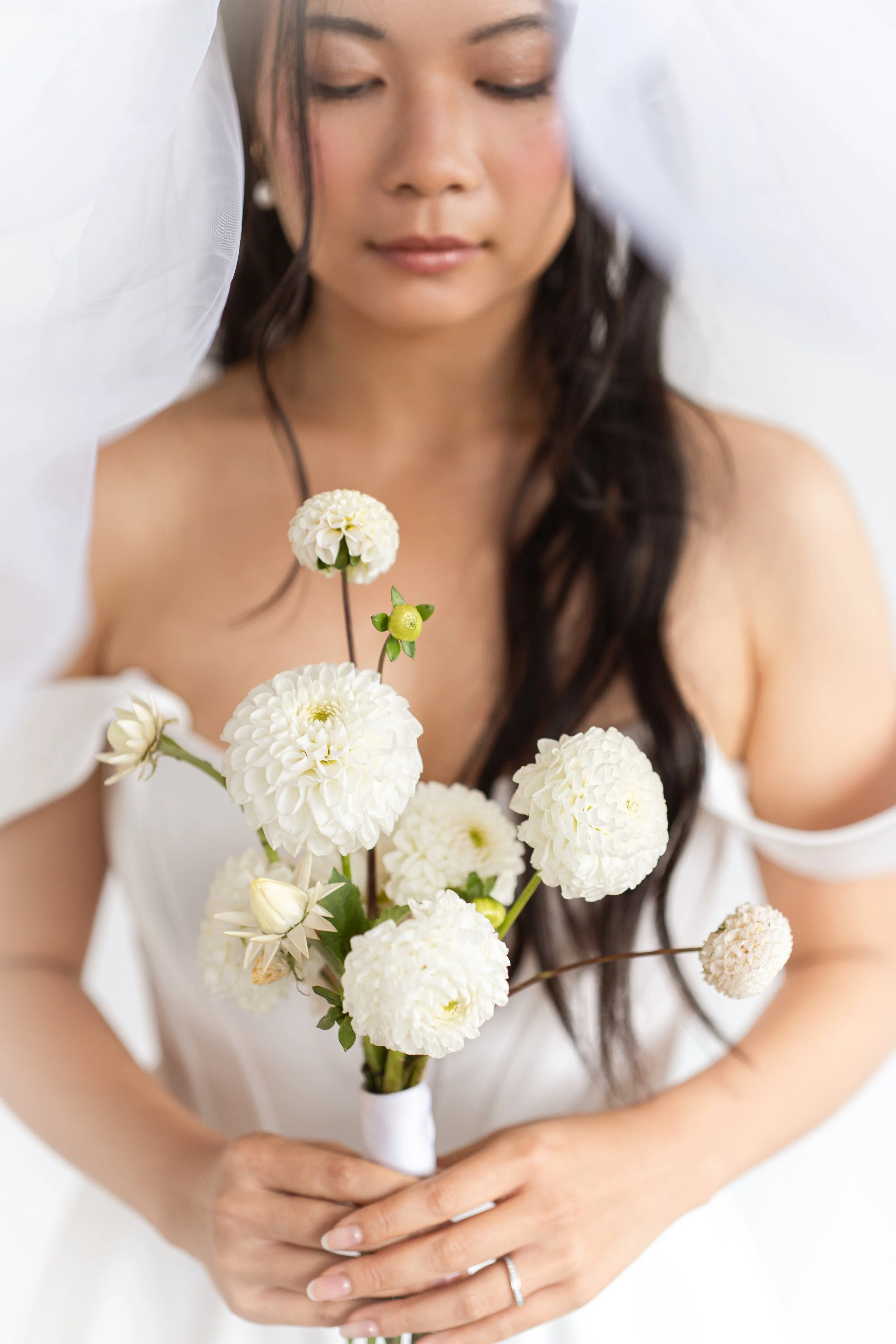 A woman with long dark hair and light skin holding a bouquet of white flowers, wearing a white off-shoulder dress, with eyes closed and a serene expression.