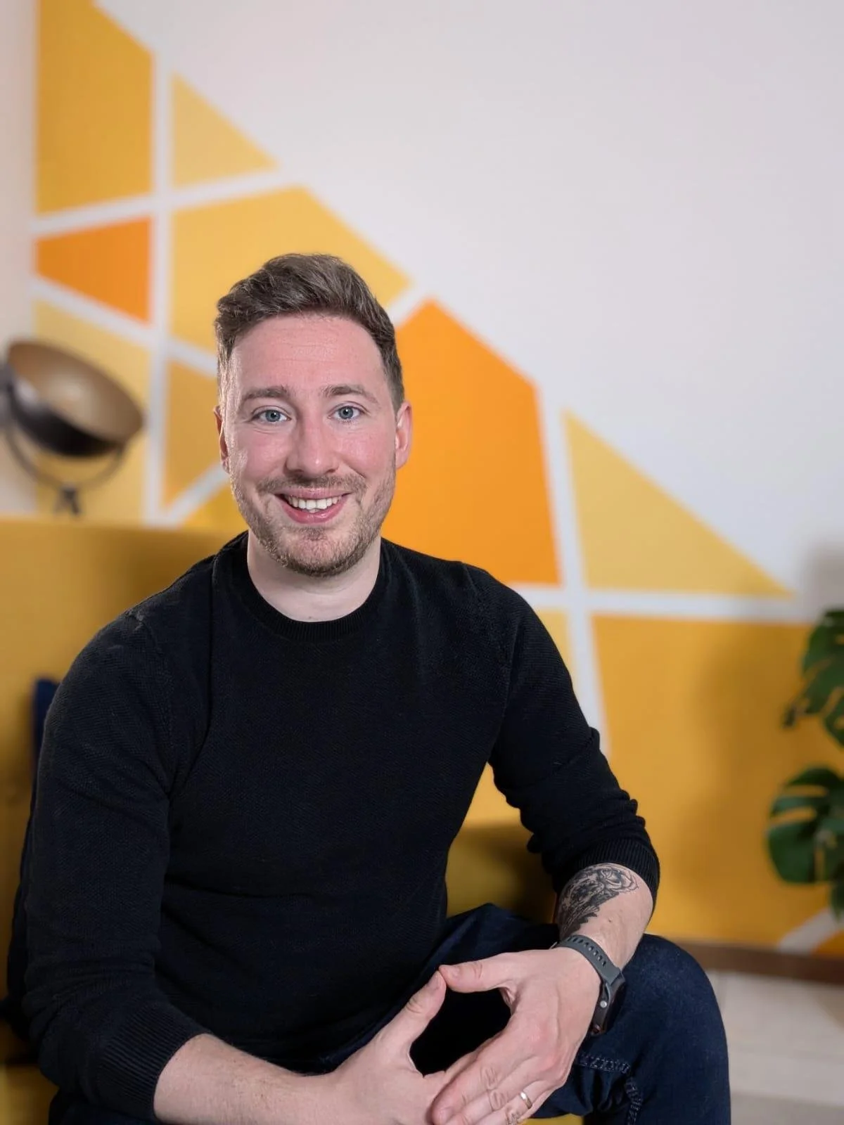 Mann mit dunklem Pullover sitzt auf gelbem Sofa vor einer Wand mit gelb-orangefarbenem geometrischem Muster.
